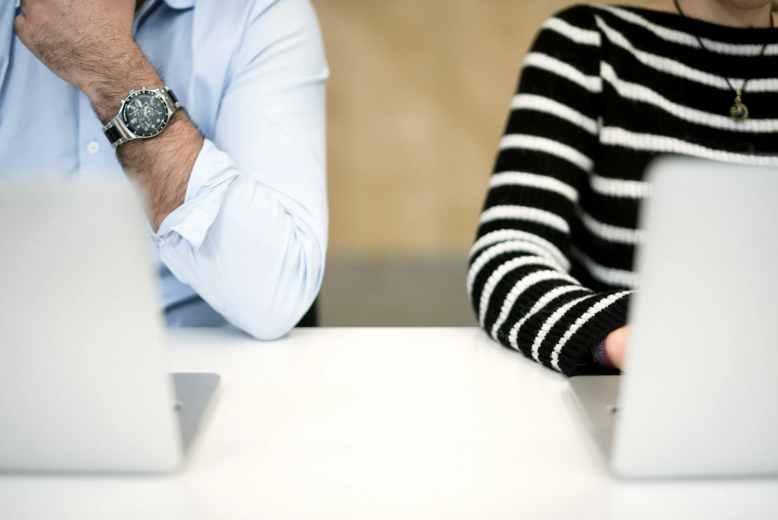 Close-up of two people sitting at a white table, one wearing a black and white striped sweater and a necklace, the other wearing a light blue shirt and a silver wristwatch.