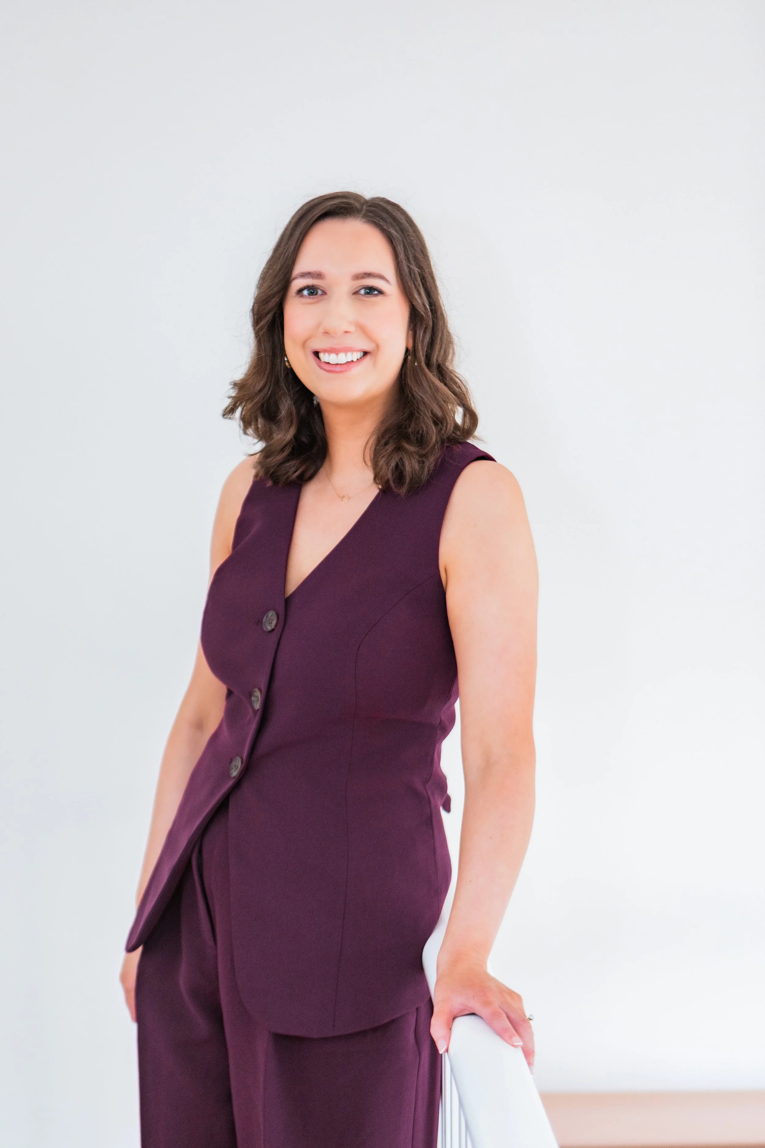 Woman called Rachel Grace Elliott with brown hair wearing a sleeveless purple dress, smiling, standing against a plain white wall, resting her hand on a white chair.