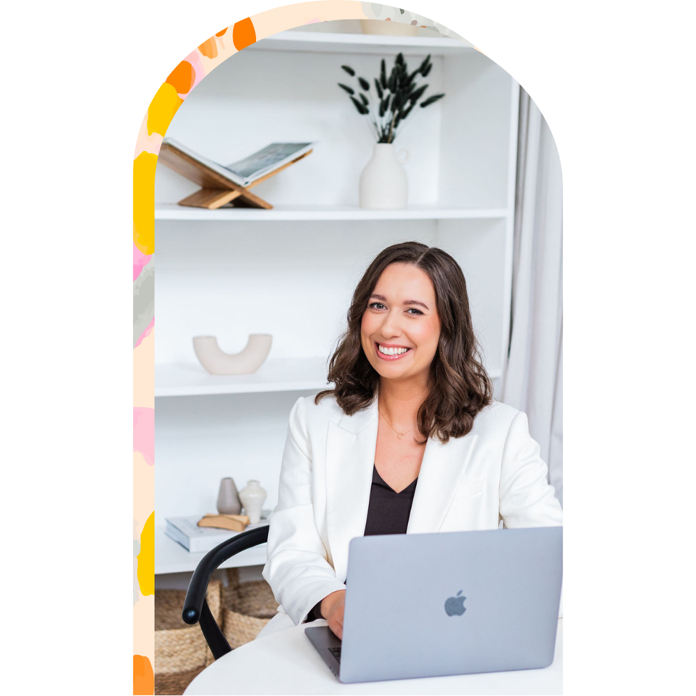 Rachel Grace Elliott smiling at a laptop in a bright, modern office with white shelves and decorative items behind her.