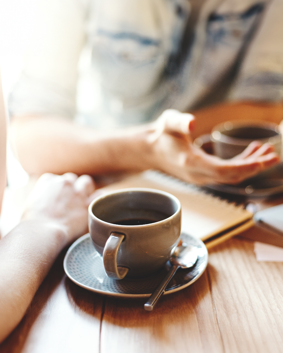 A cup of black coffee on a saucer with a spoon, on a wooden table, with a person's arm and hand gesturing in the background.