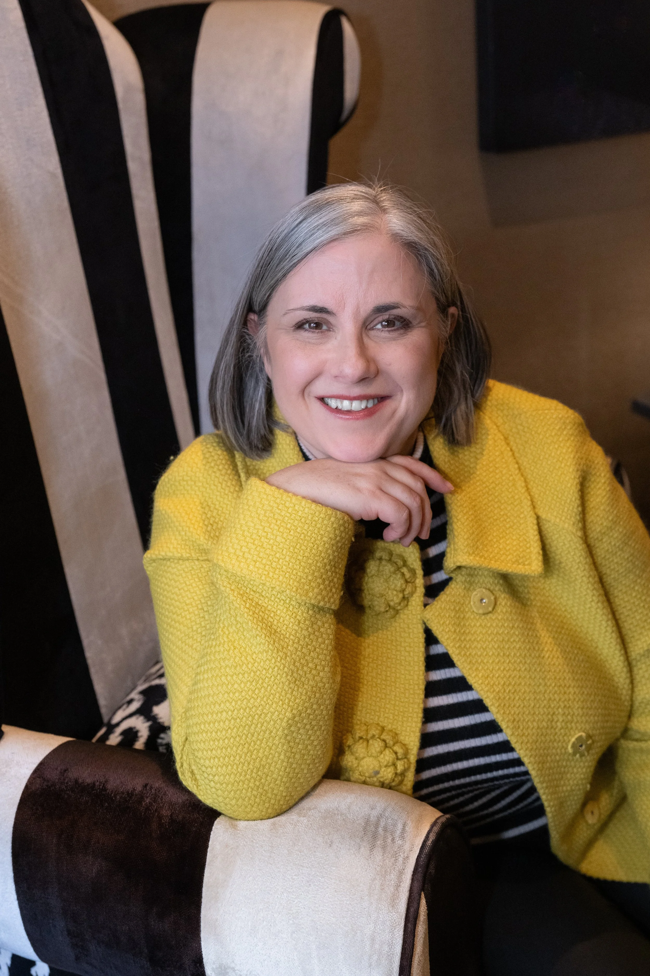 Coach Jo - A woman with grey hair, smiling and wearing a yellow textured jacket, sitting on a patterned upholstered chair.