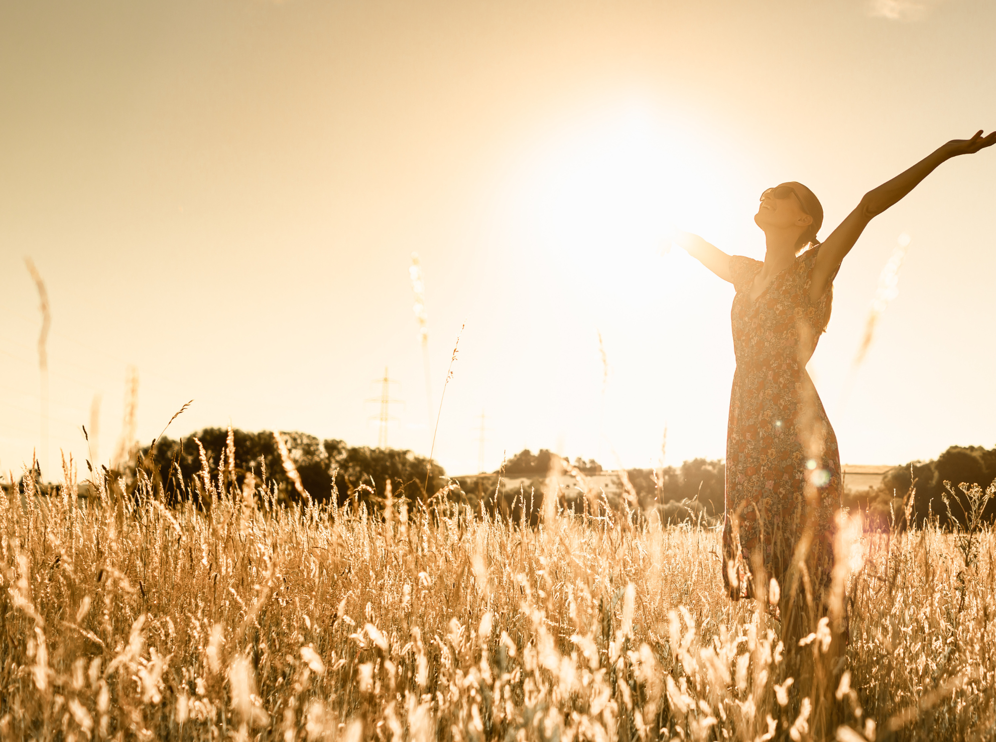 A woman with arms outstretched standing in a golden wheat field during sunset, smiling with the sun illuminating her face and body.
