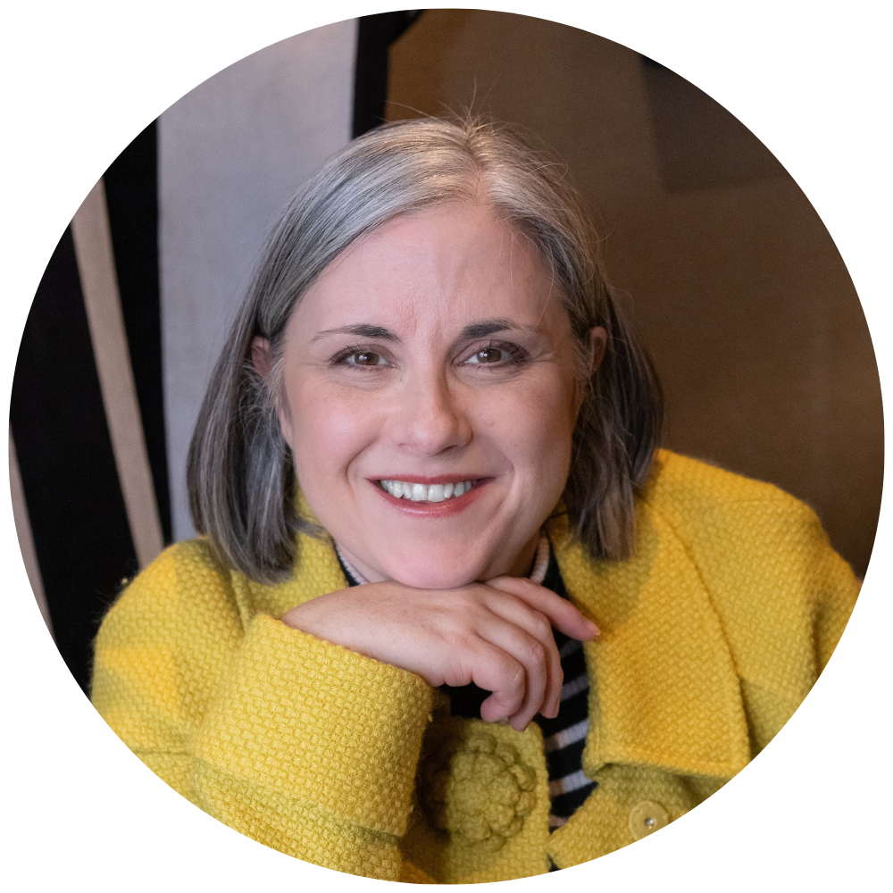 Coach Jo - A woman with grey hair, smiling and wearing a yellow textured jacket, sitting on a patterned upholstered chair.