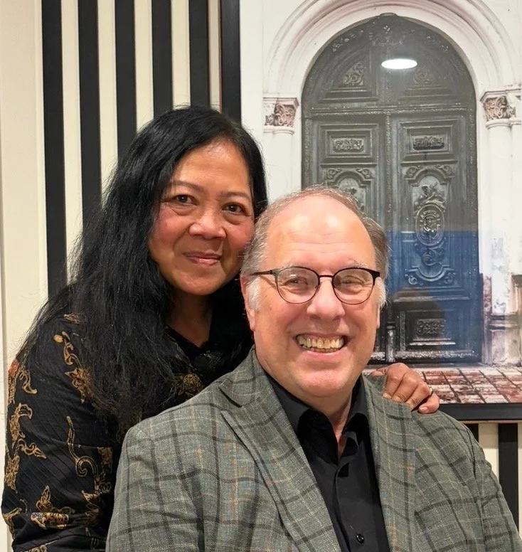 A woman with long black hair and a man with short gray hair and glasses are posing together, smiling indoors in front of a backdrop depicting a large ornate door.