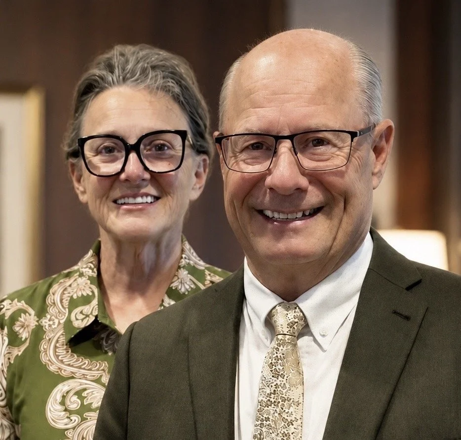 A smiling elderly man and woman, both wearing glasses, in formal attire. The man is in a suit with a patterned tie, and the woman is wearing a green patterned blouse. They are standing indoors with warm lighting.