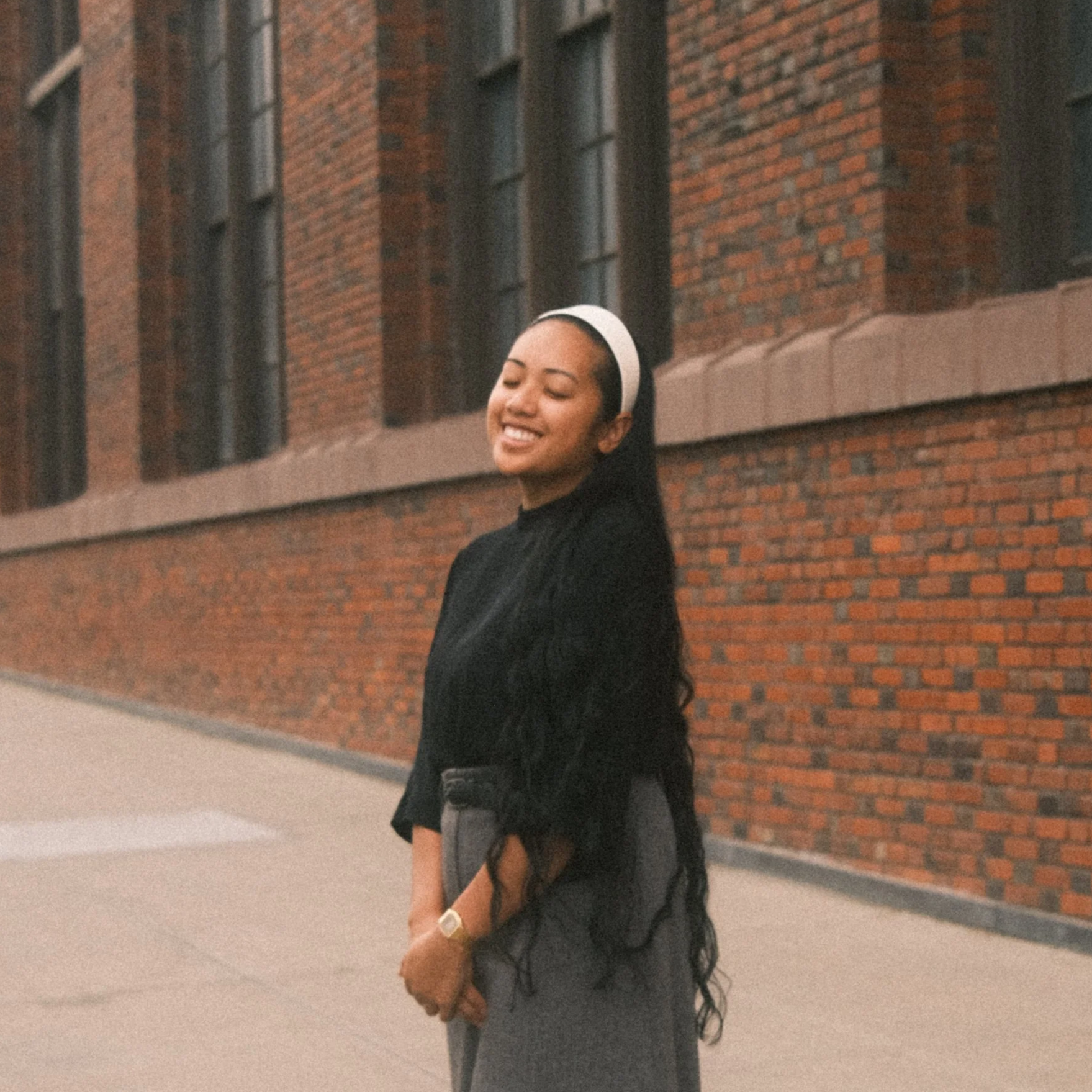 Young woman with long dark curly hair wearing a white headband, black blazer, and gray pants, smiling with eyes closed standing outdoors in front of a brick building.