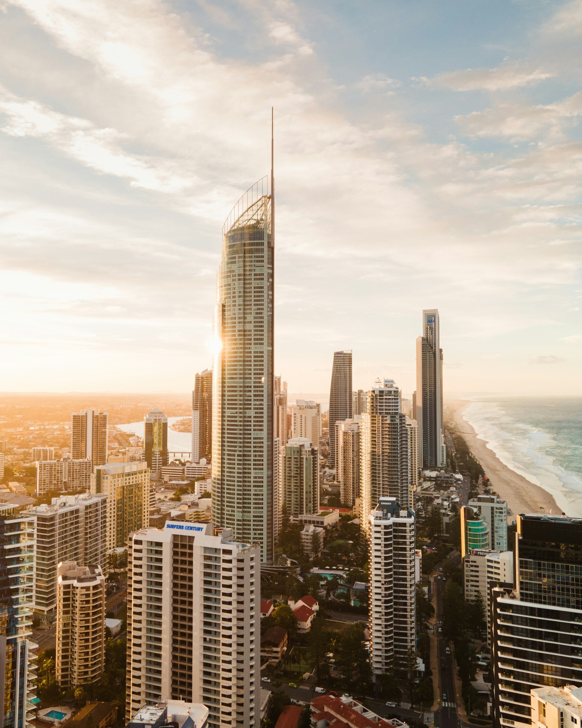 A city skyline with high-rise buildings at sunset, overlooking a beach and ocean to the right, with a partly cloudy sky.