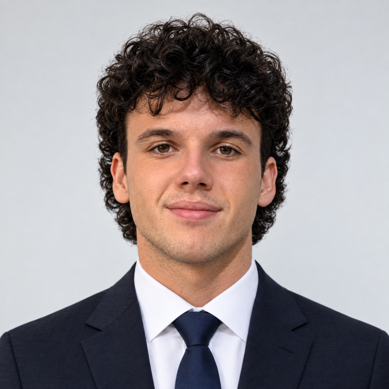 Young man with curly dark hair in a navy suit and tie, smiling mildly, against a plain light background.