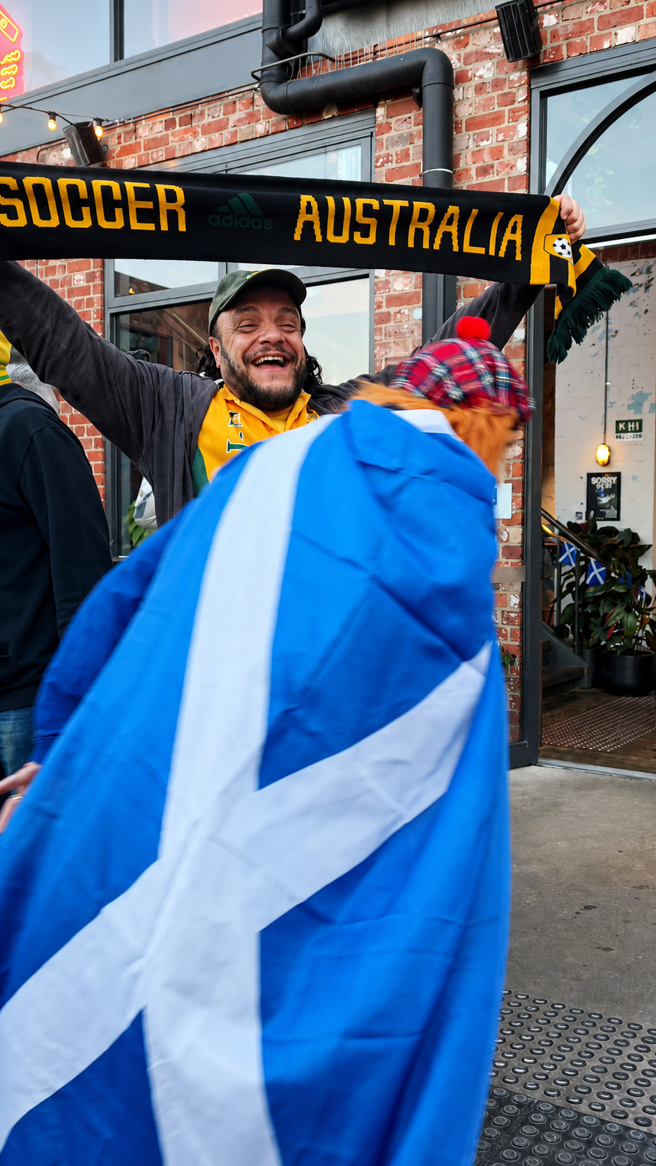 A man smiling and celebrating while holding an Australia soccer scarf in front of a brick building, with a person draped in a Scottish flag in the foreground, outside a stadium or sports venue.