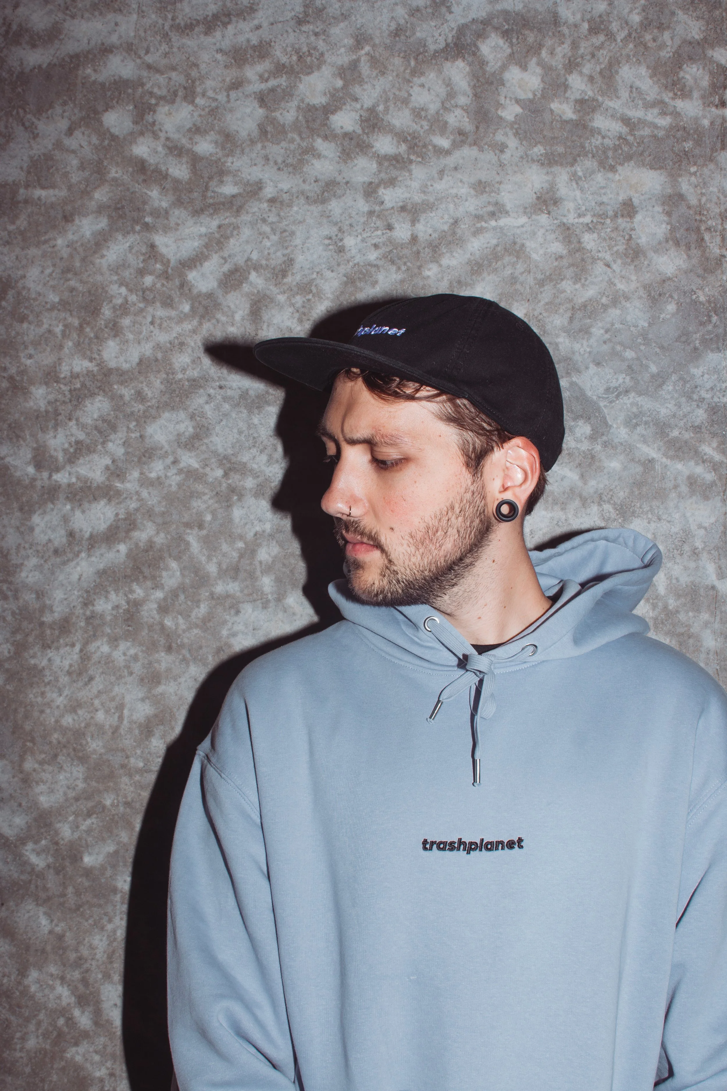 A young man with a beard and stretched earlobe piercing, wearing a black cap and light blue hoodie, standing against a textured gray wall, with eyes closed and head slightly tilted downward.