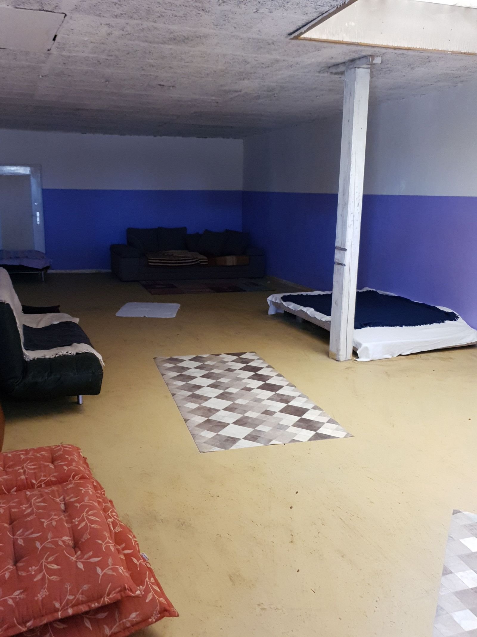 Ein Raum mit einfacher Einrichtung, blauer Wand, einem Sofa, einem Bett, einem Teppich und einer Trennwand.