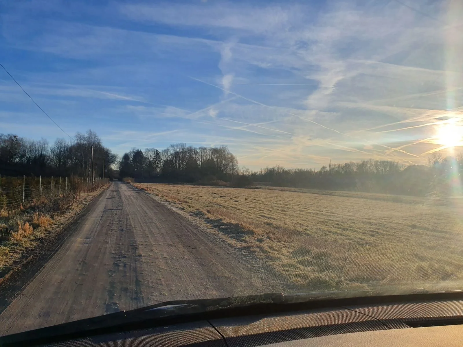 Eine unbefestigte Landstraße in einer ländlichen Gegend, umgeben von Feldern und Bäumen, bei Sonnenuntergang mit einem blauen Himmel und einigen Wolken.