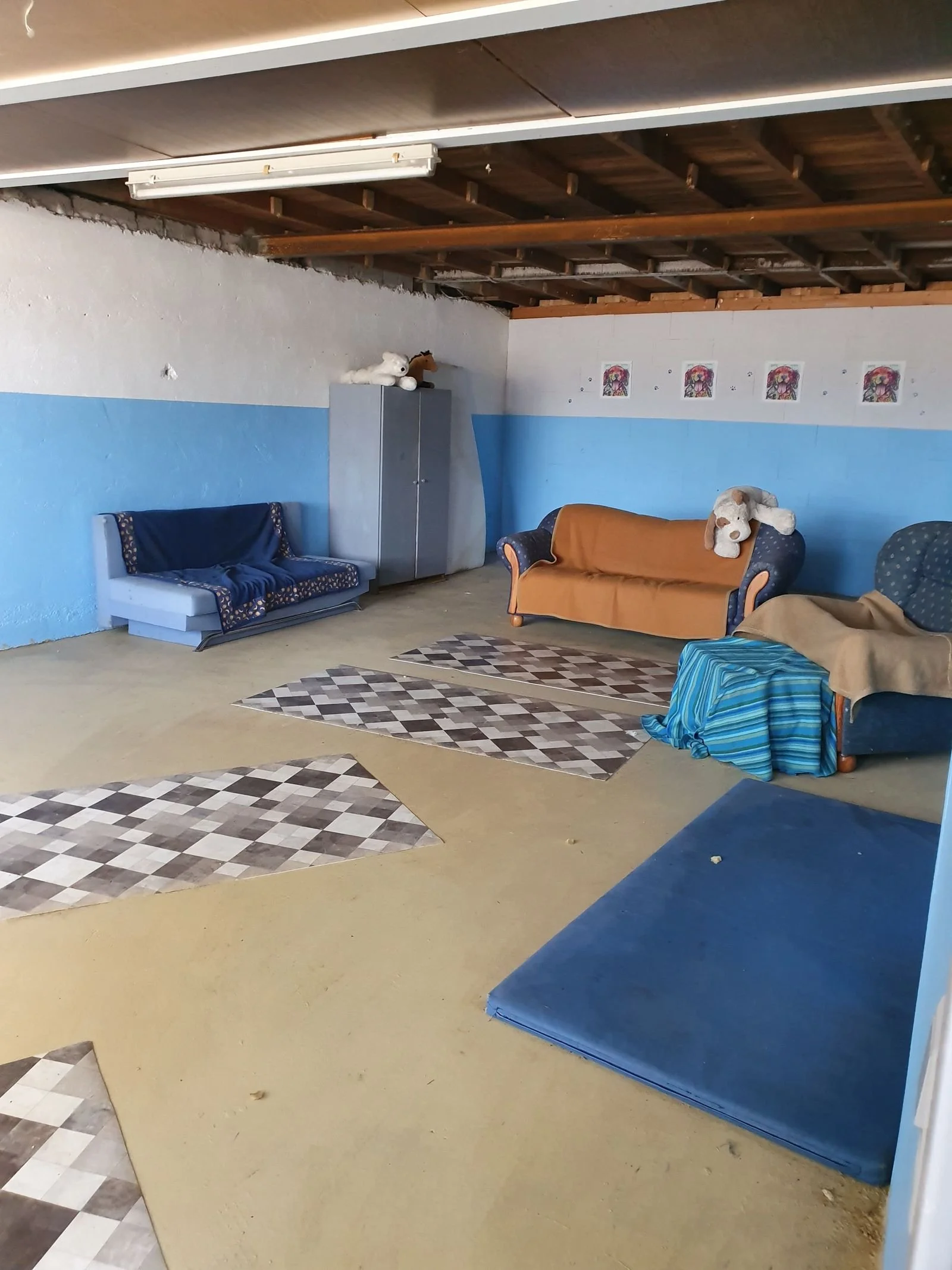 Ein Raum mit blauen Wänden, Sofa und Teppichen, die auf einem gelben Boden liegen. An der Wand befinden sich kleine Bilder, in einer Ecke steht ein Schrank mit Stofftieren auf der oberen Ablage.