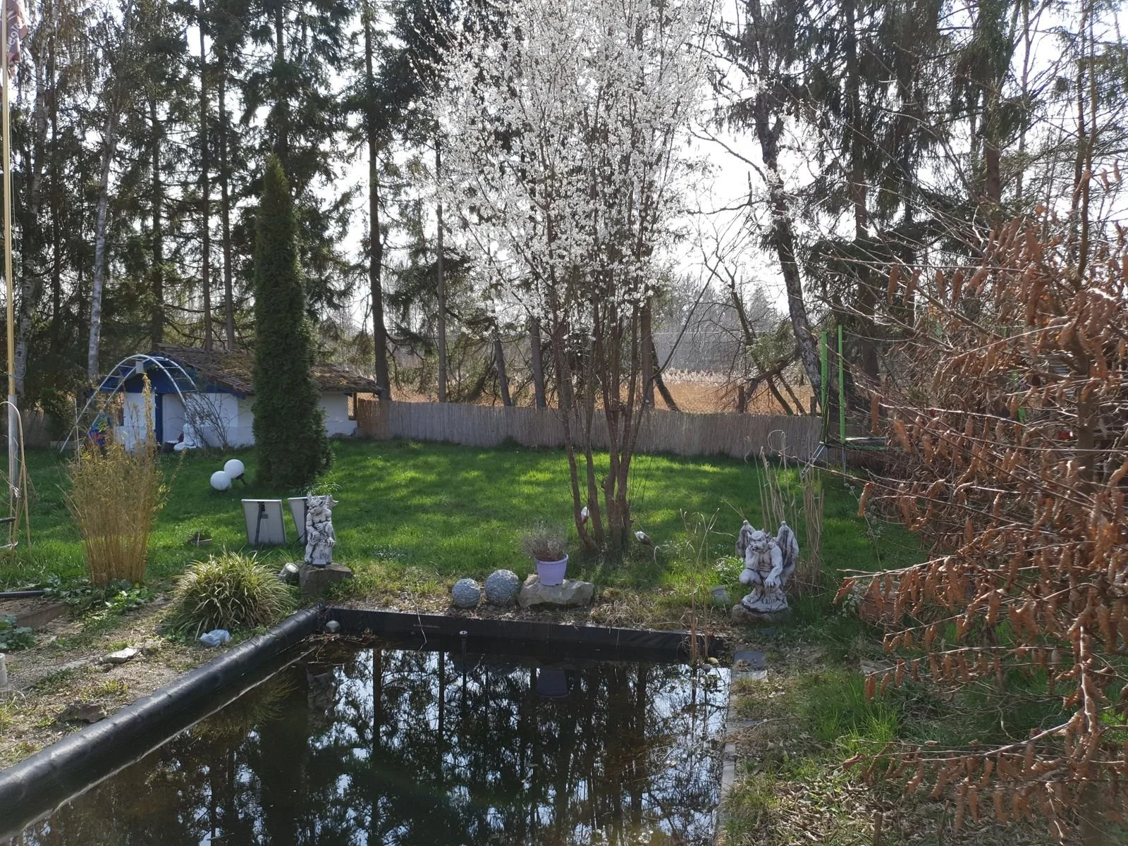 Garten mit Wasserbecken, Steinskulpturen, Bäume und Büsche, Zaun und kleine Hütte im Hintergrund, sonniger Tag.