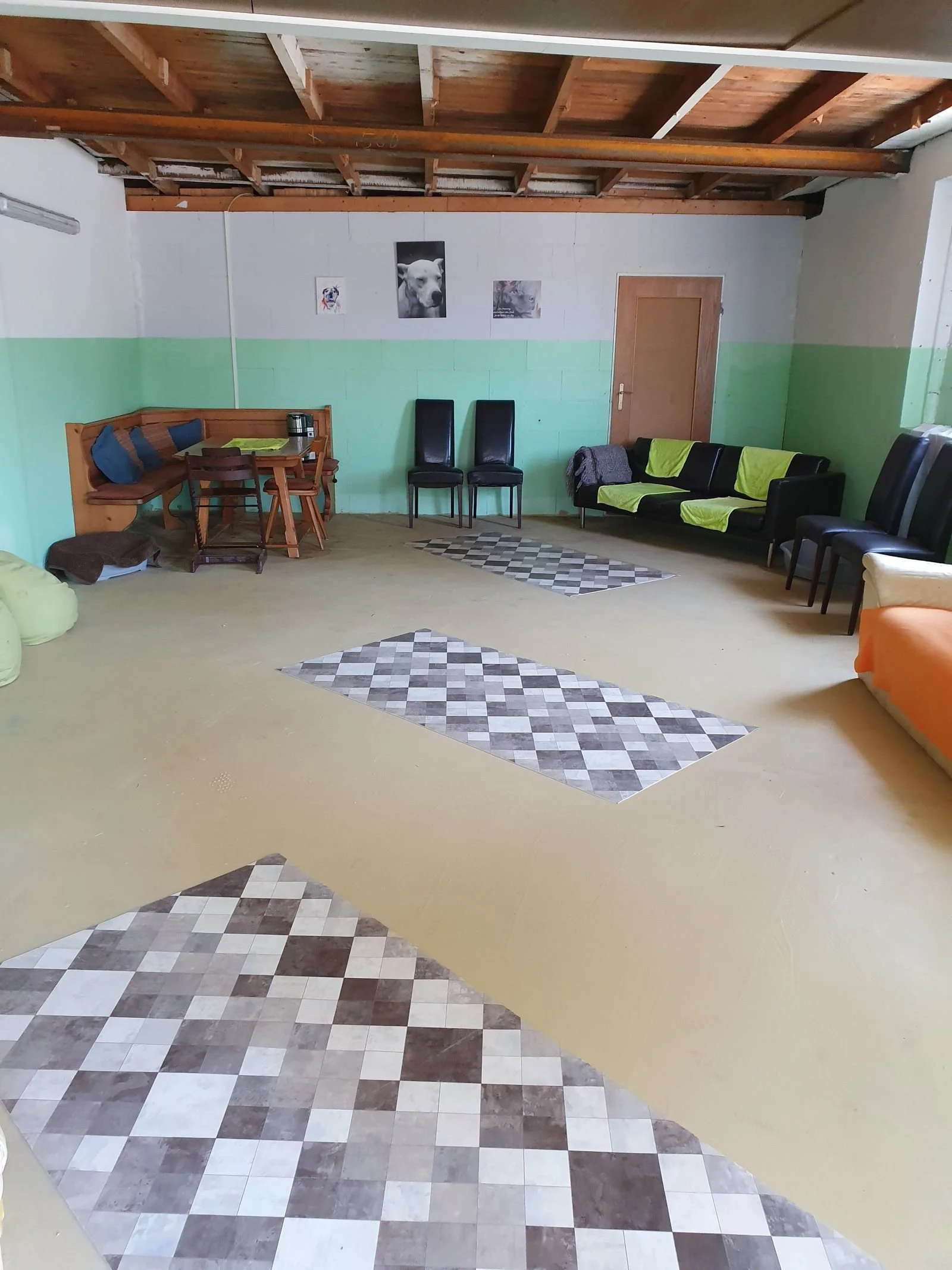 Ein Raum mit gemusterten Bodenfliesen, einer grünen Wand und mehreren schwarzen Stühlen, einer Couch mit grünem Bezug, Wanddekorationen mit Tierbildern, Holzdecke mit sichtbaren Balken und einem Holzhaustür.