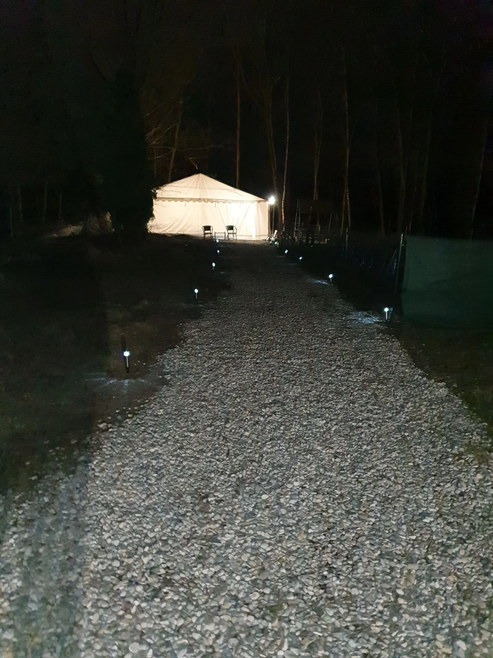 Ein unbeleuchteter Weg auf einem Campingplatz, mit kleinen Steinen bedeckt, geführt zu einem weißen Zelt und zwei Stühlen darin, umgeben von dunklen Bäumen bei Nacht.