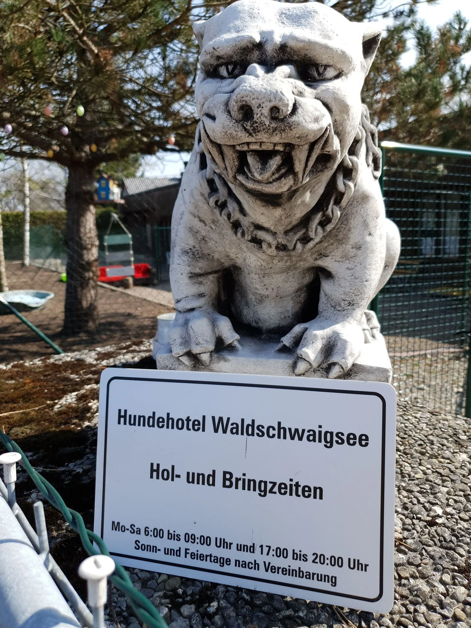 Steinfigur eines Löwen vor einem Baum mit bunten Kugeln, daneben ein Schild mit Öffnungszeiten für das Hundehotel Waldschwaigsee.