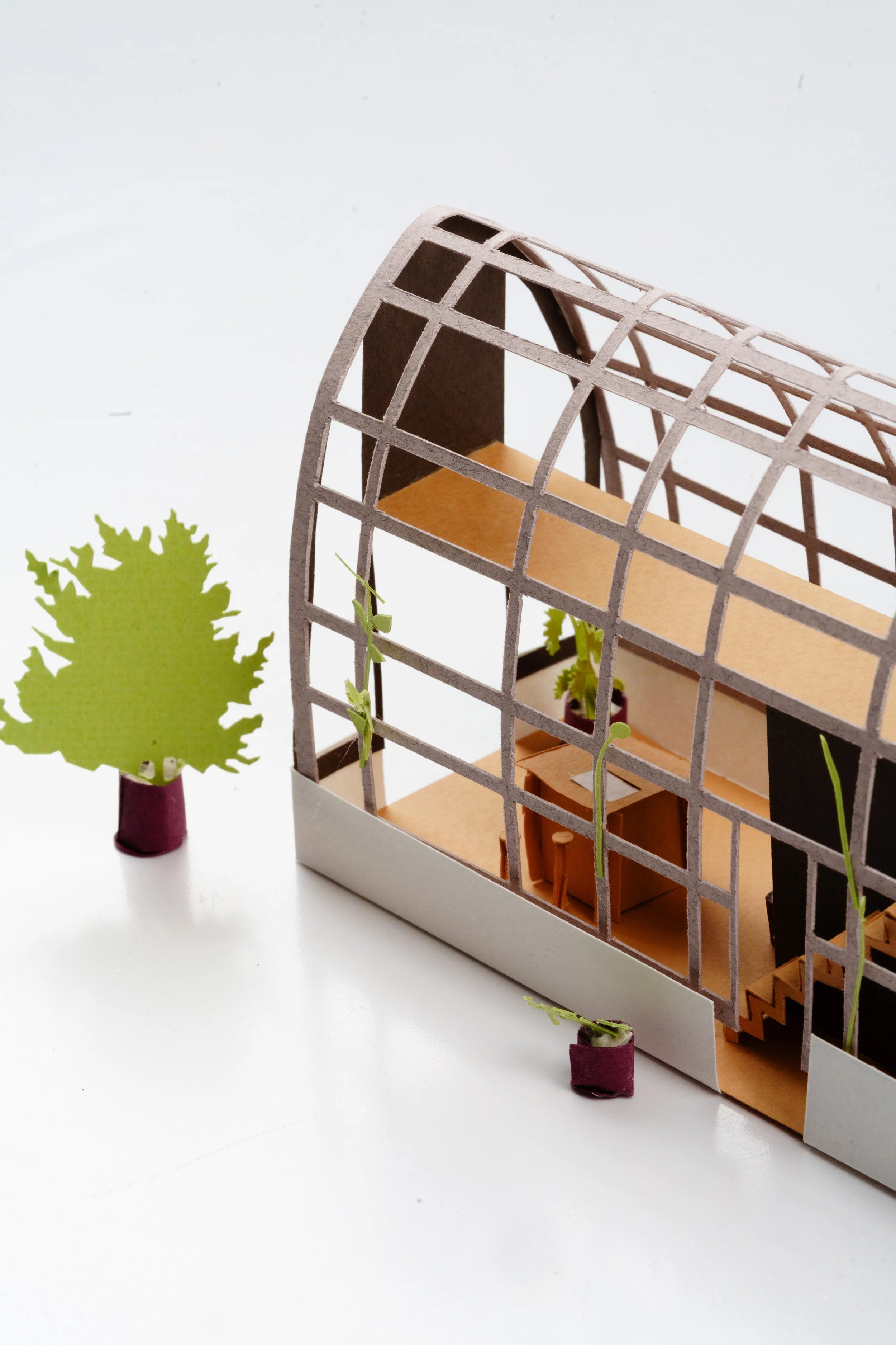 A miniature architectural model featuring a modern, curved, steel grid-like structure with an open interior and a wooden bench inside. There are small potted plants with green leaves and purple pots to the side.