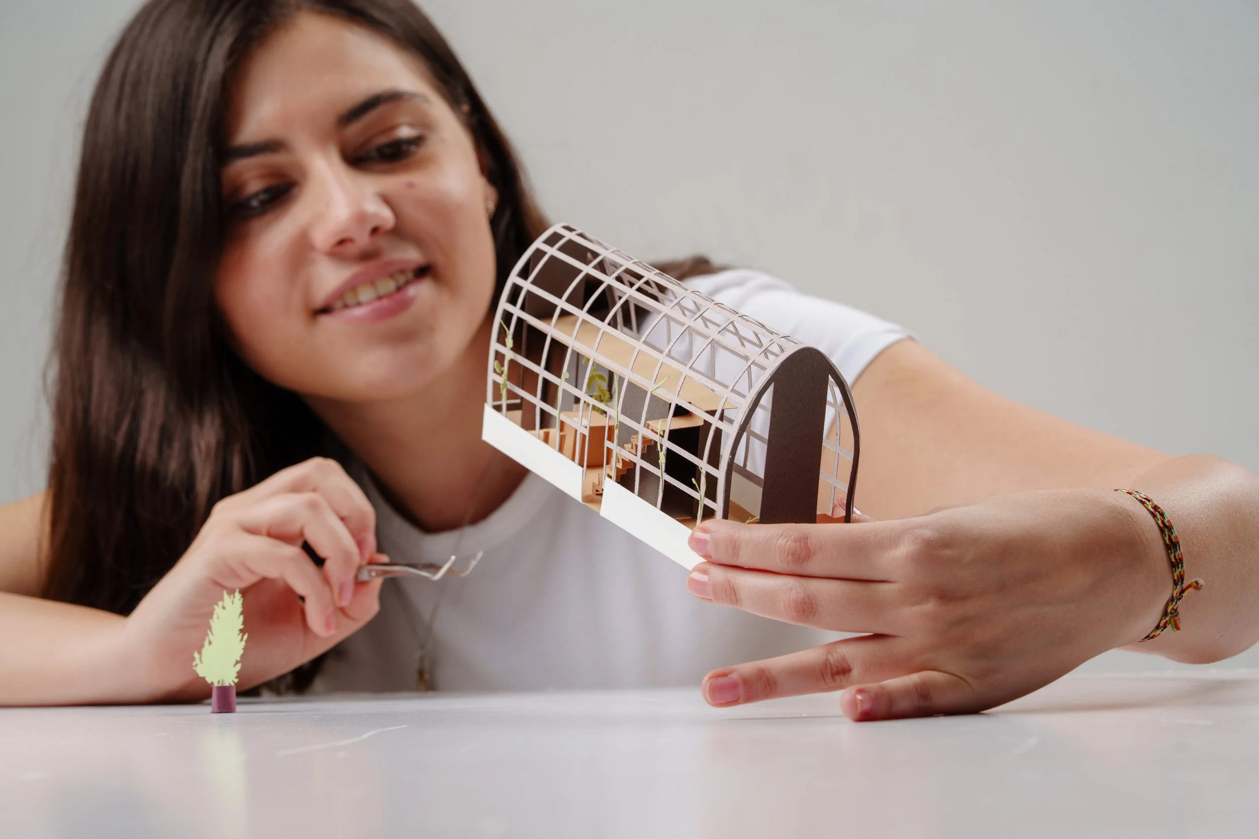 A young woman with long brown hair is holding and looking at an architectural model of a building with a rounded, open framework design. She is sitting at a table with her arm resting on it, and there is a small, lit, paper tree plant on the table. Her expression is pleasant and focused.