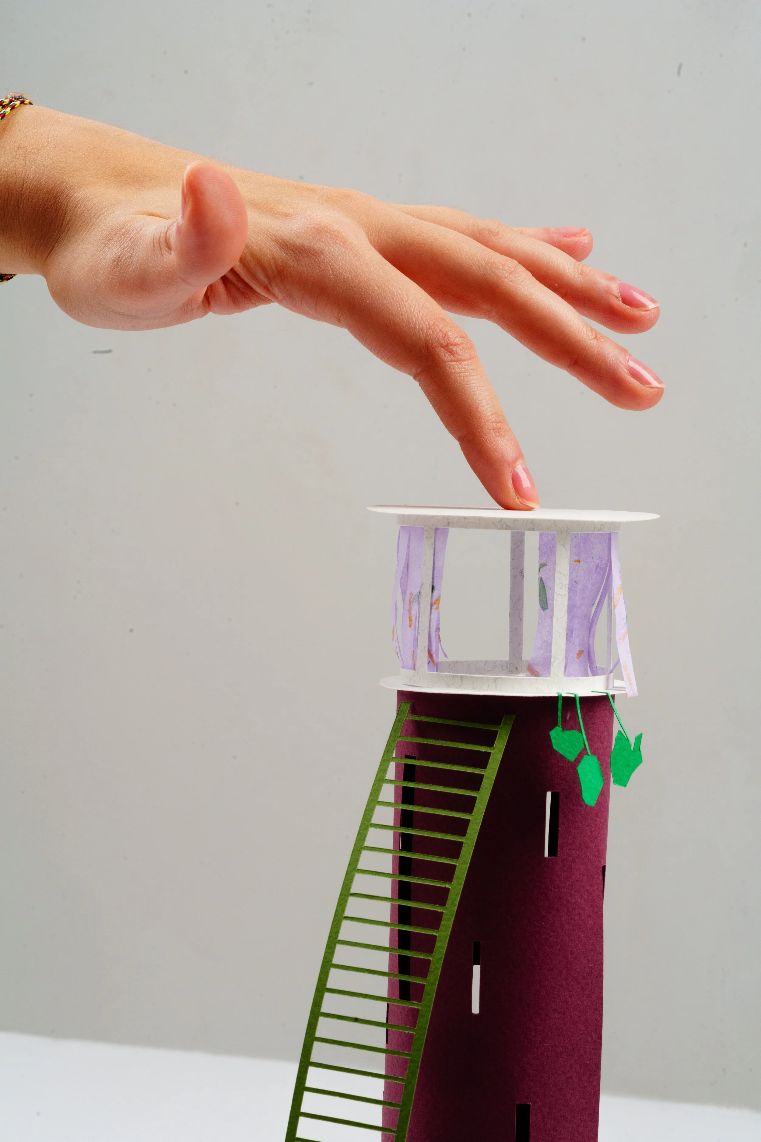 A craft project of a multi-story tower made from paper, with a hand placing a paper piece on the top. The tower has a pink base with a staircase cutout and green paper trees hanging from it, and a purple midsection with a ladder cutout. The background is plain and off-white.