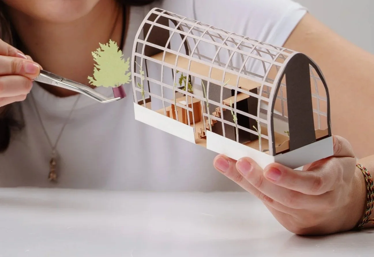 Person holding a small architectural model of a greenhouse or botanical conservatory, with green paper or plastic plants inside, using tweezers to place a small plant on the model.
