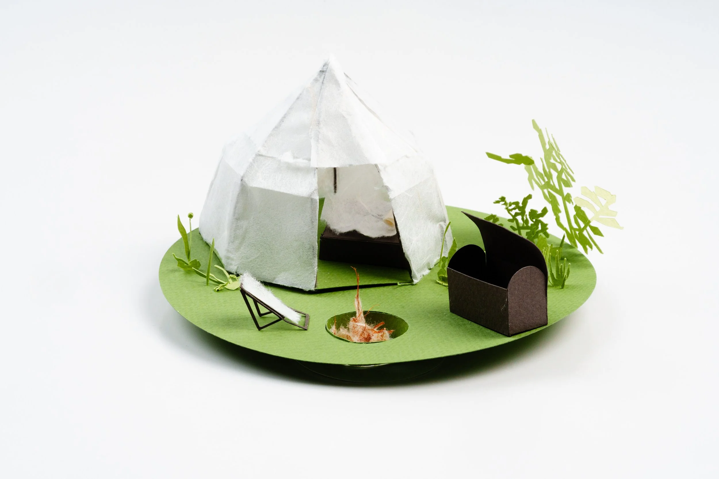 A miniature model of a teepee on a green circular base with plants, a small black box, and a tiny footbridge.