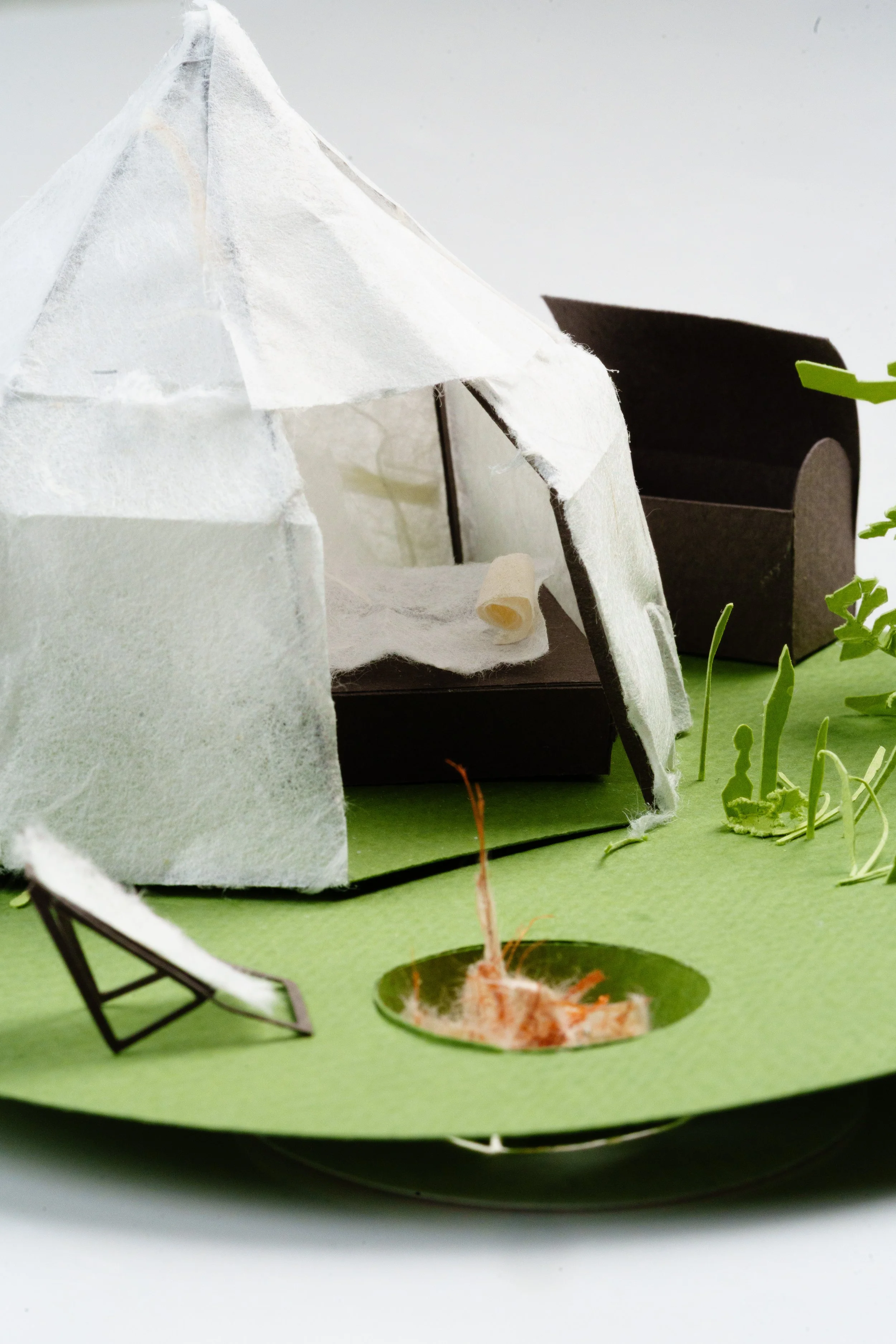 A miniature diorama featuring a small black structure, white textured rocks, a tiny chair, a small garden bed with tomato plant, and a hole in the ground with visible root.