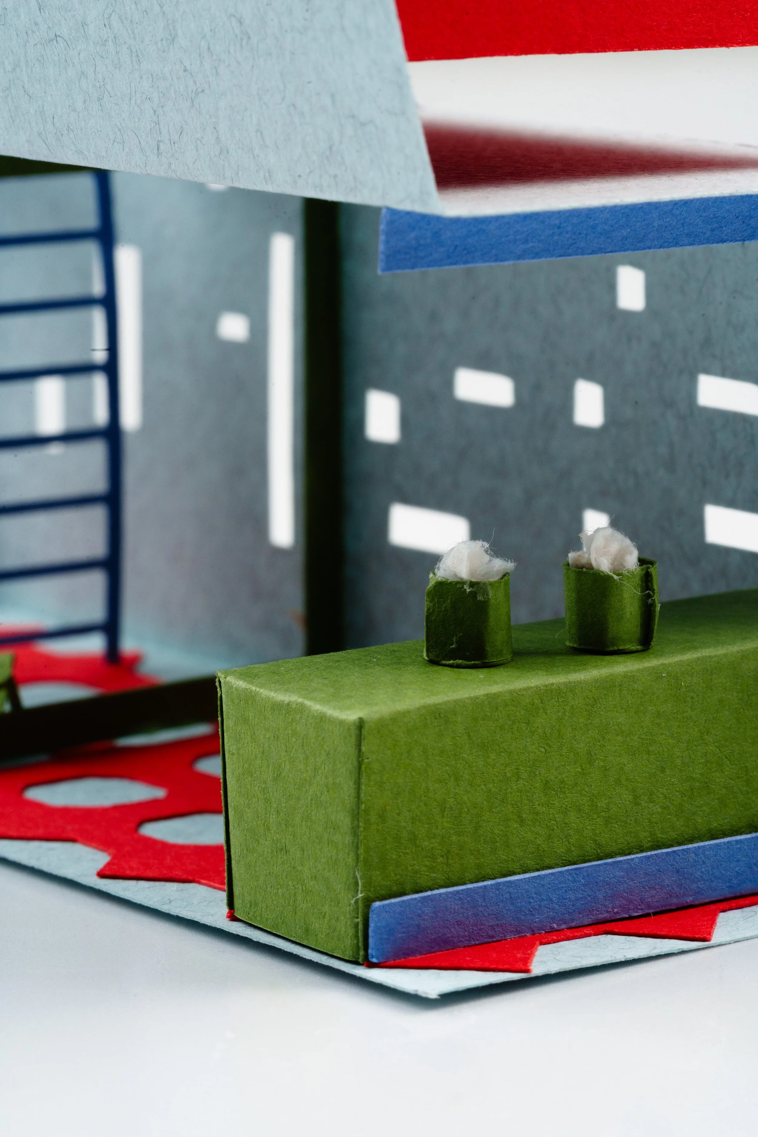 Close-up of a colorful, abstract paper craft scene with a green paper block, small paper cylinders with cotton at the top, and a background with blue, red, and white paper layers with cut-out patterns.
