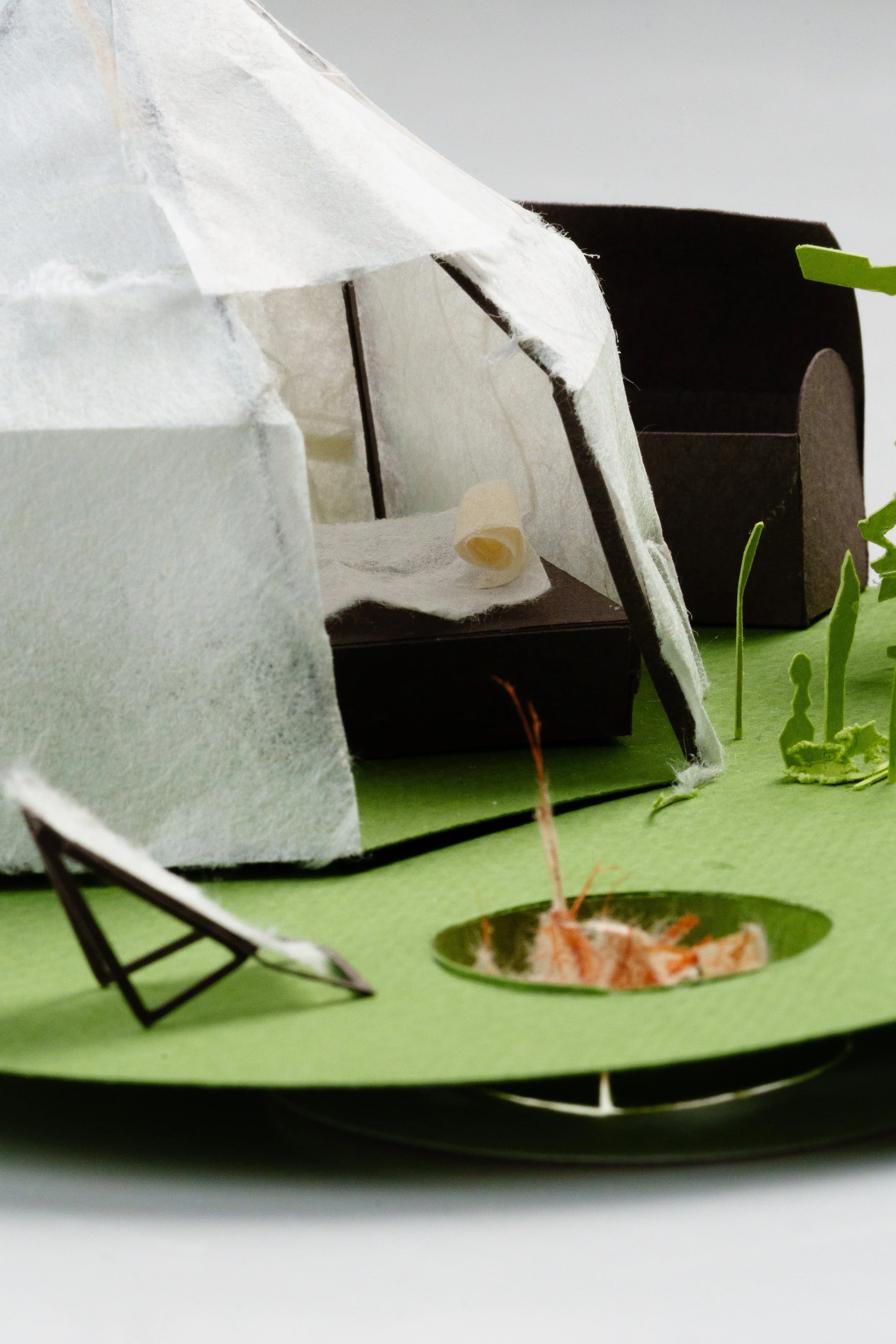 Miniature model of a house with grey walls and a triangular roof, outdoor area with green grass, small plants, and a round opening with shredded paper inside, situated on a black circular base.