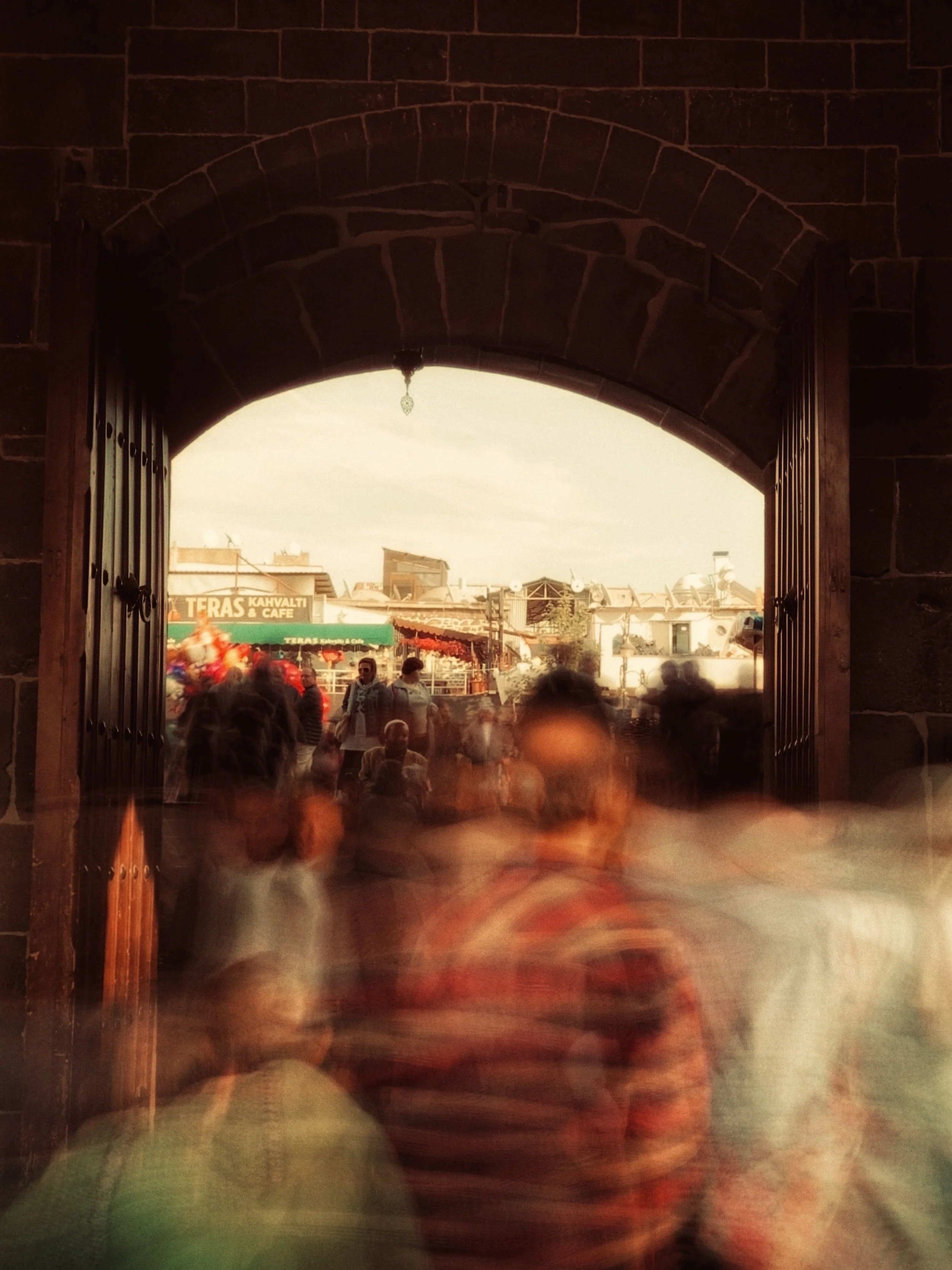 People inside a building looking through an archway to an outdoor scene with buildings and a sign that reads 'TERAS KAHVALTI & CAFE.'