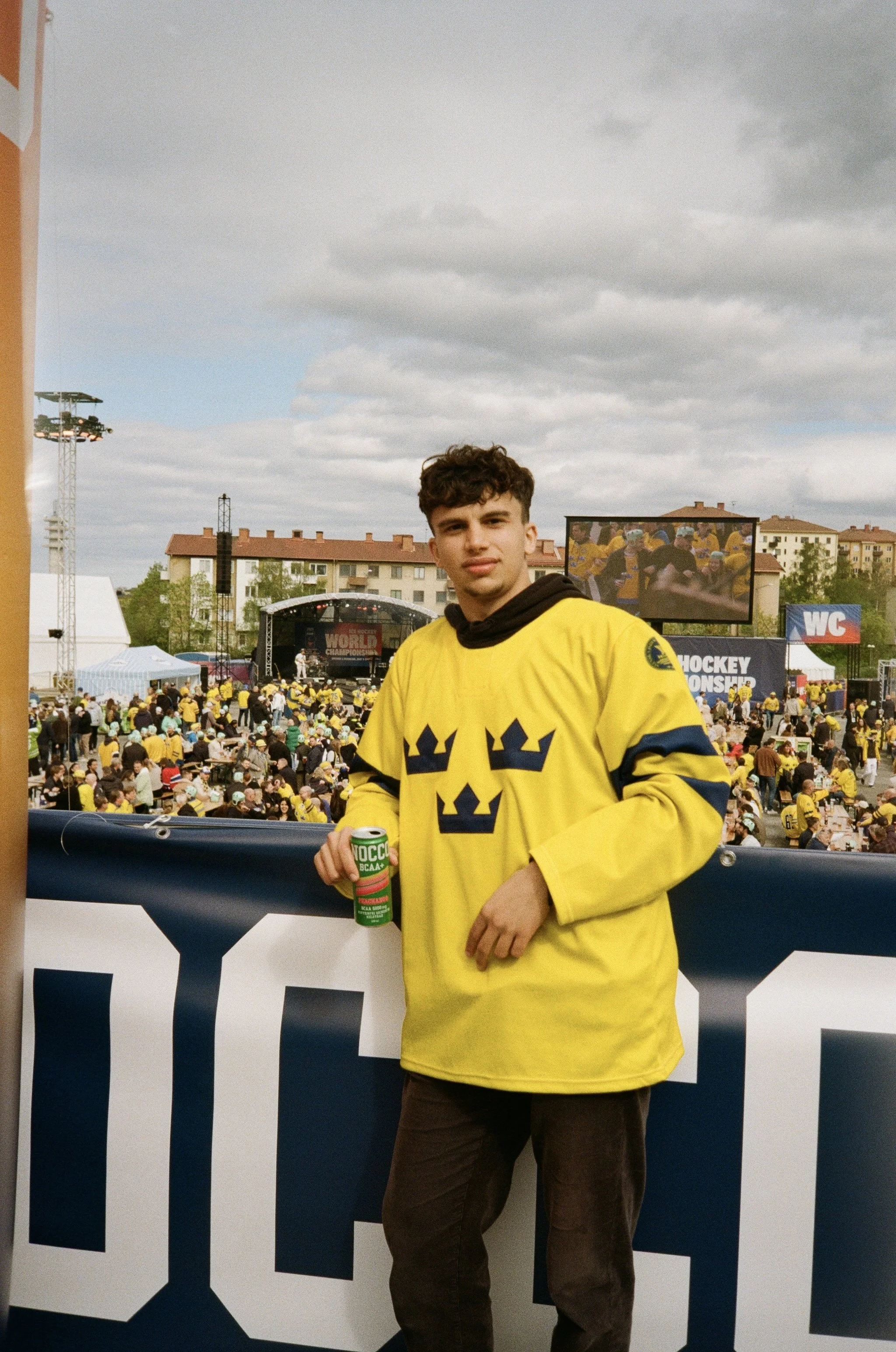 Young man wearing a yellow hockey jersey holding a can of energy drink at an outdoor event with a crowd and large screen in the background.