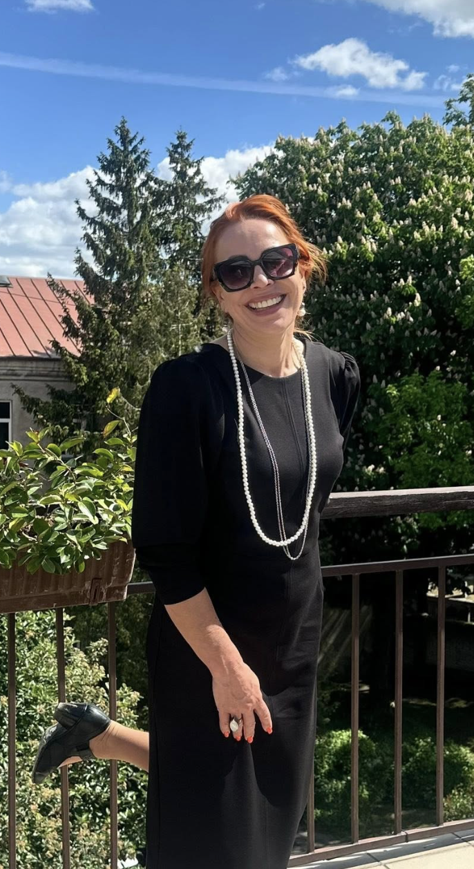 A woman with red hair and large sunglasses smiling outdoors on a sunny day, wearing a black dress, a pearl necklace, and black shoes, standing on a balcony with greenery and trees behind her.