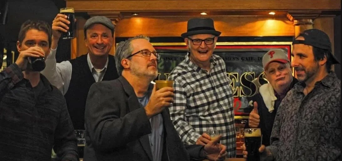 Four men in a pub holding dark beers and smiling for a group photo.