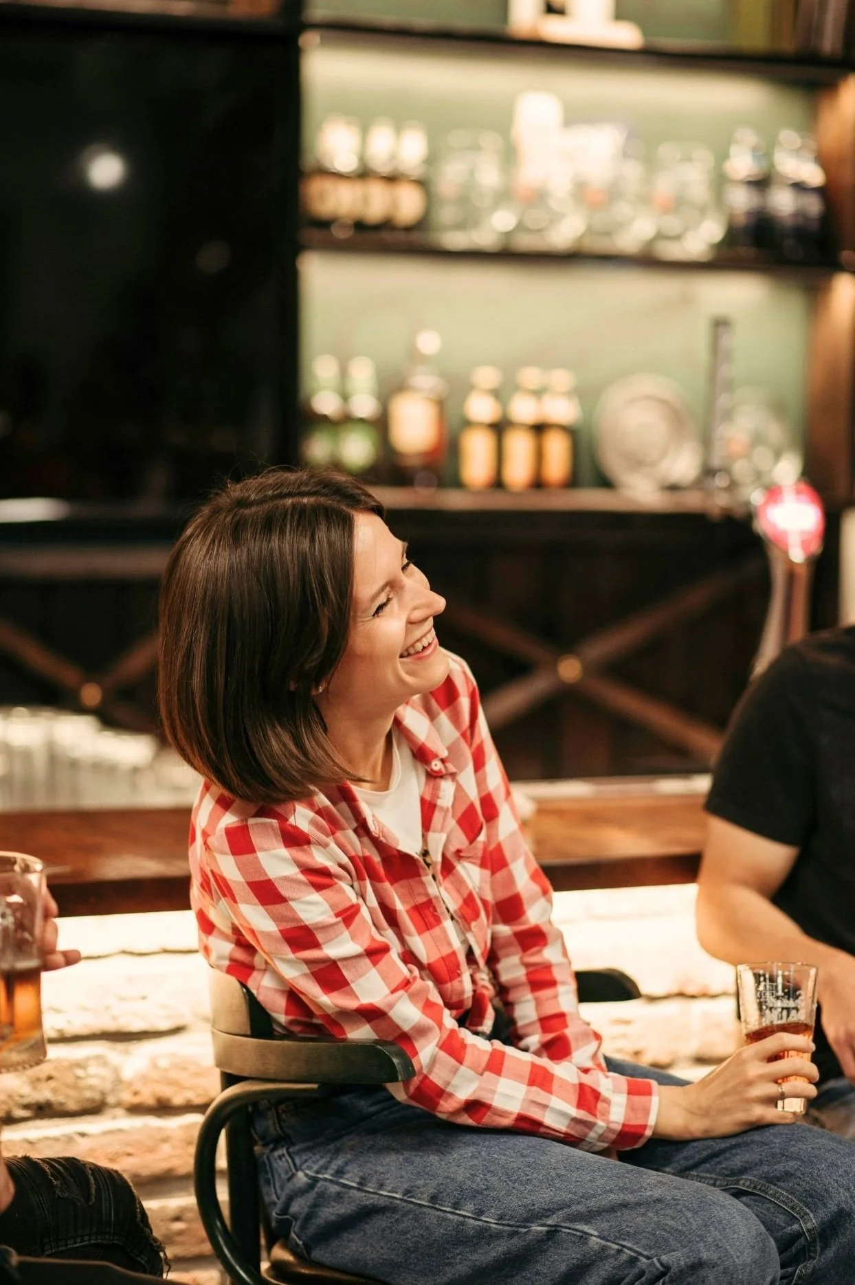 Woman in a red plaid shirt smiling while chatting at a bar.