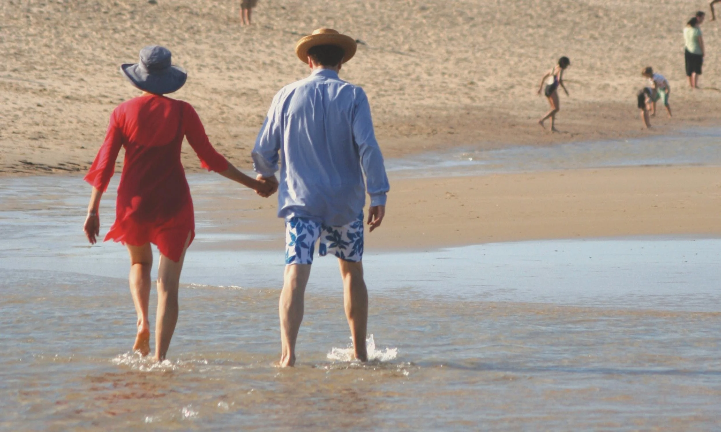 Older couple holding hands while walking along a sandy beach shoreline.