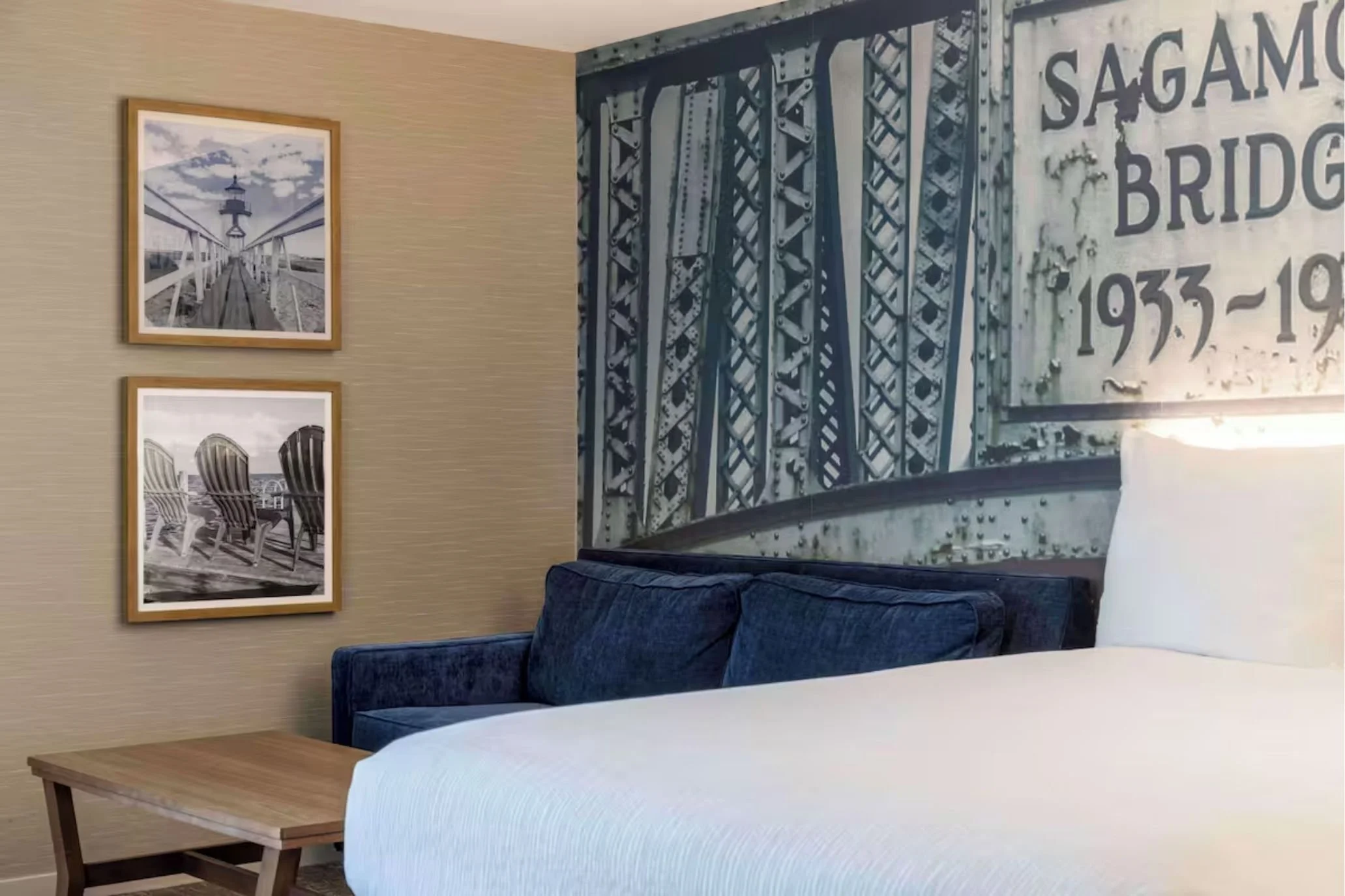 Cozy hotel suite with a bed, blue loveseat, coffee table, and decorative bridge mural backdrop.