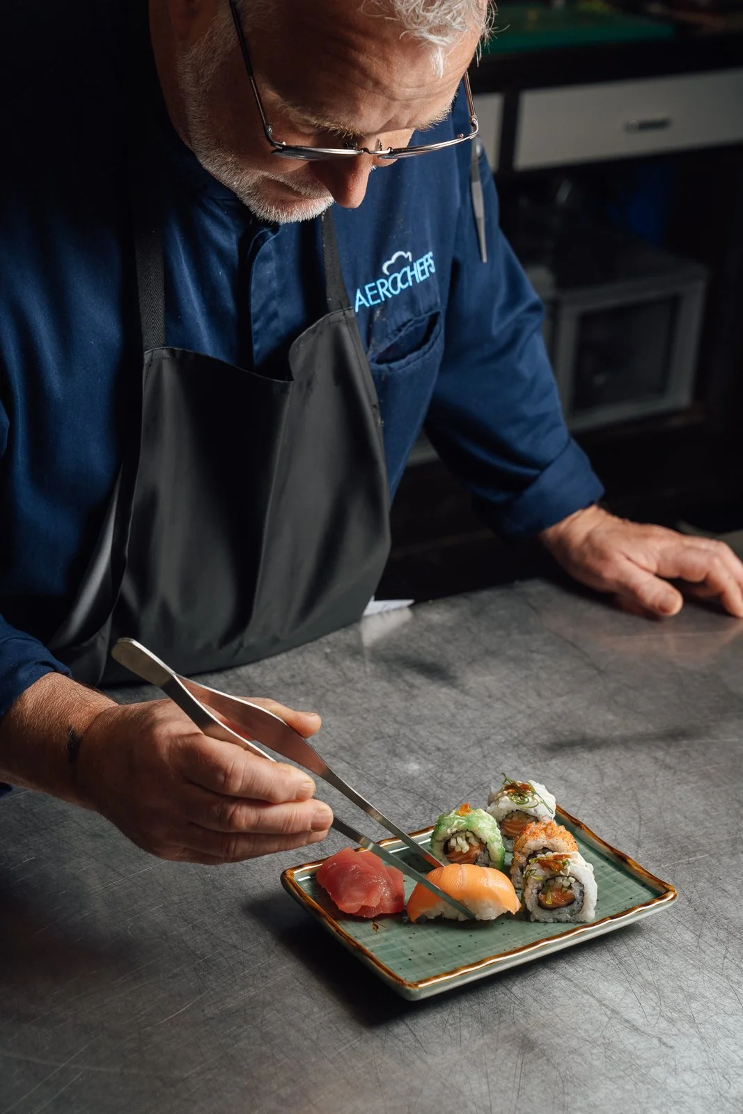 Een chef-kok bereidt sushi op een blauw bord, hij gebruikt een tang om de sushi te plaatsen. De chef draagt een smal zwart schort en een donkerblauw overhemd met logo.