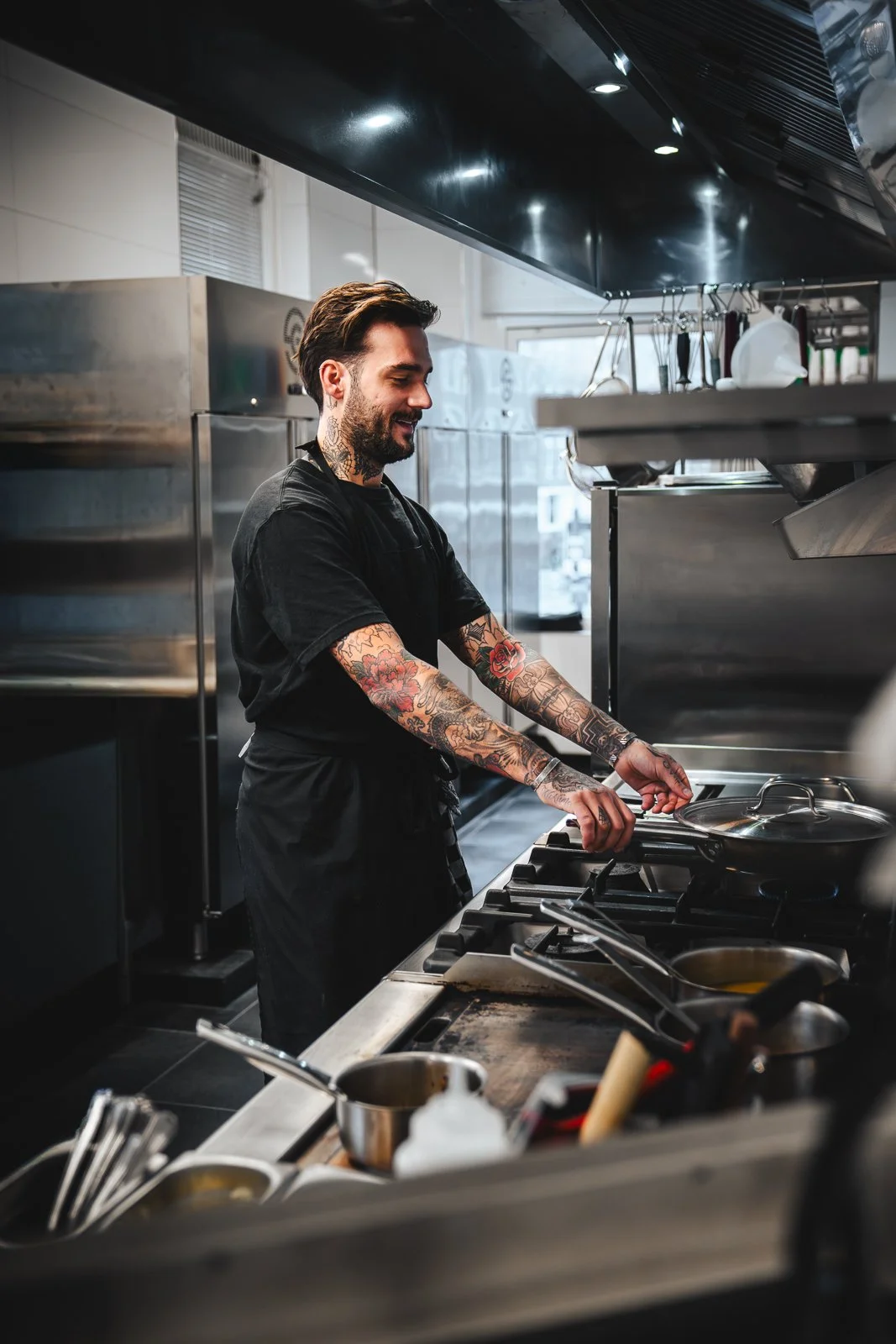 Kok in een professionele keuken, rokend vanaf een pan op het fornuis, met tattoos op armen, professionele keukenapparatuur op de achtergrond.