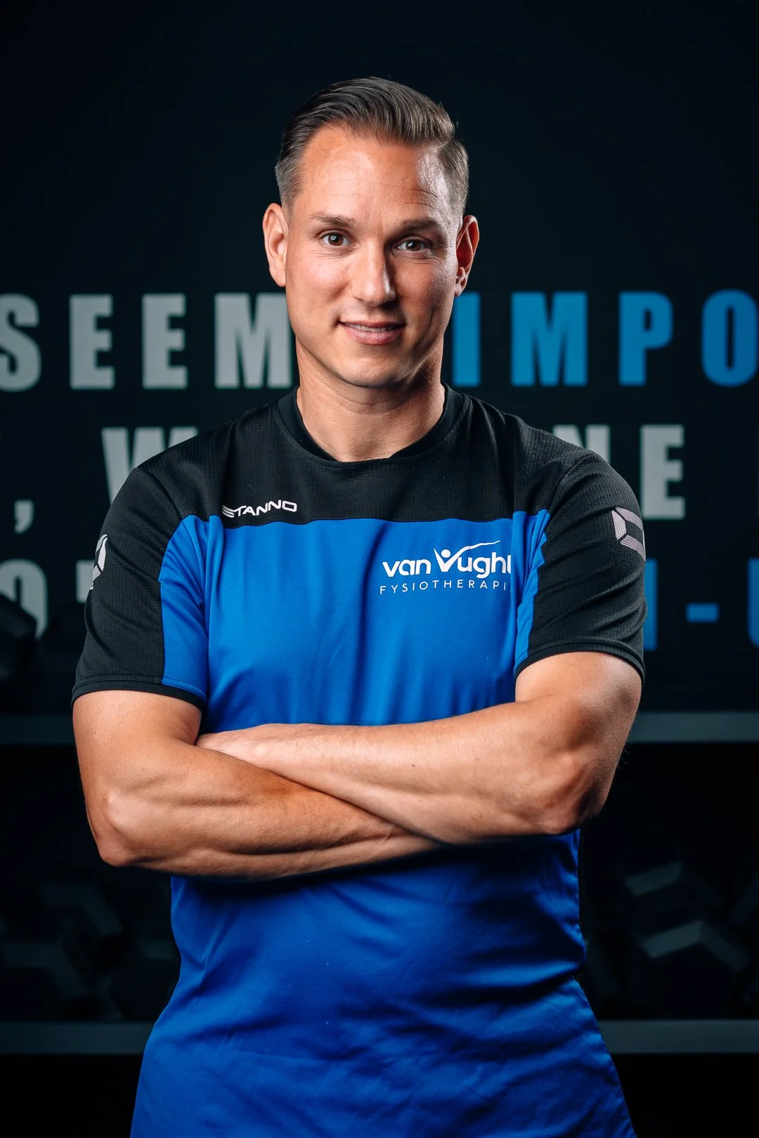Man in sportshirt met gekruiste armen, blauwe en zwarte kleuren, met tekst 'vanVugh' op de voorkant, staand voor een donkere achtergrond met tekst.