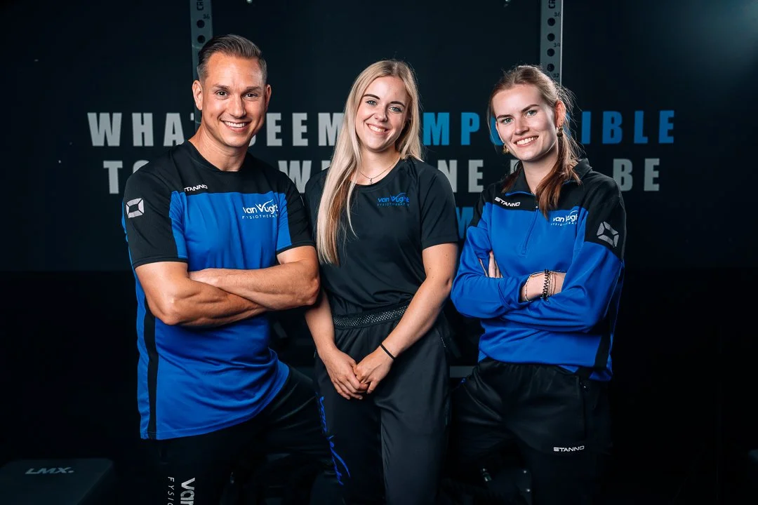Drie mensen poseerden samen, twee vrouwen en een man, in sportkleding bij een sportschool of fitnesscentrum, met een donkere achtergrond en teksten op de muur.