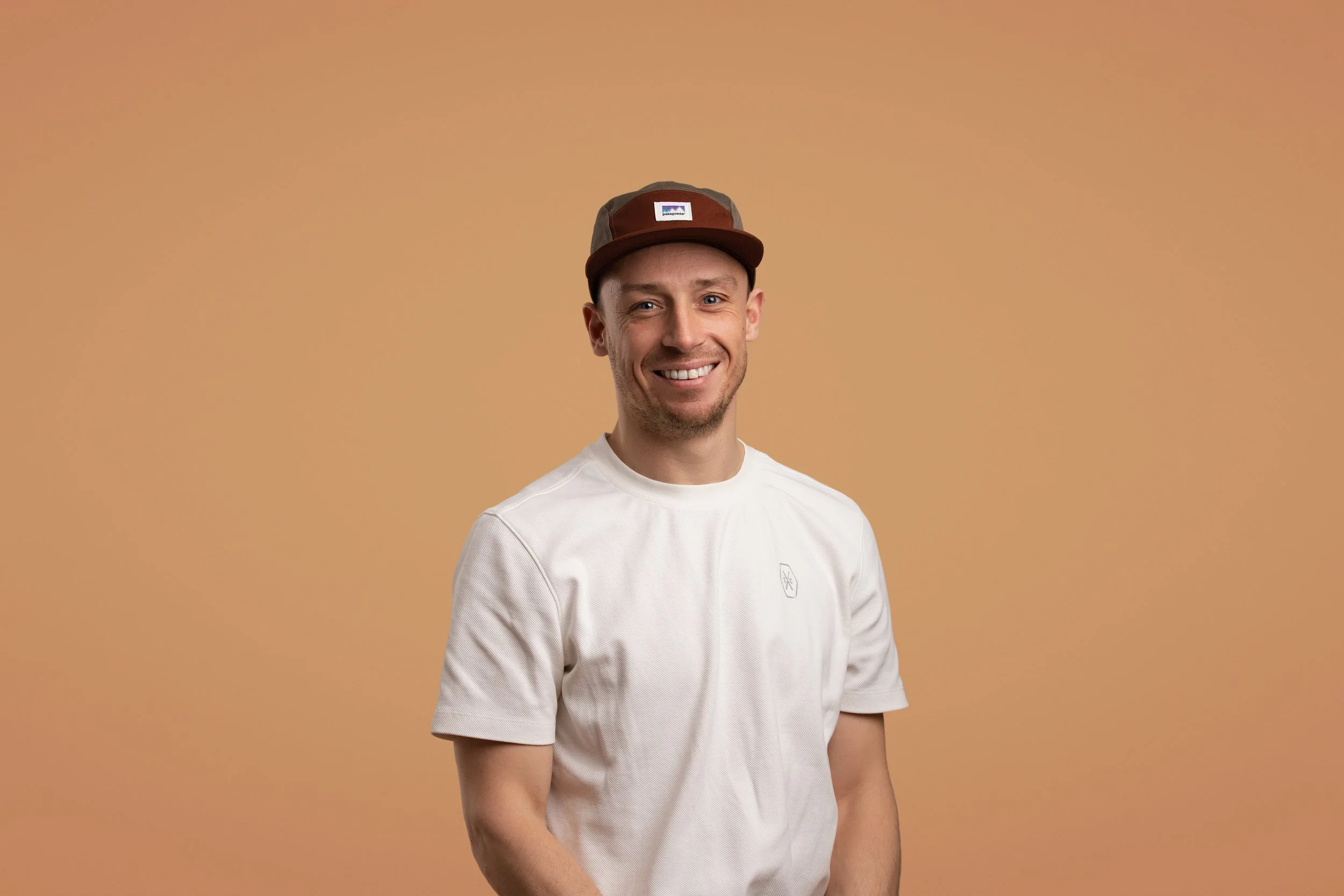 Een man met een lichte huidskleur draagt een bruin petje en een witte T-shirt, lachend tegen een lichtbruine achtergrond.