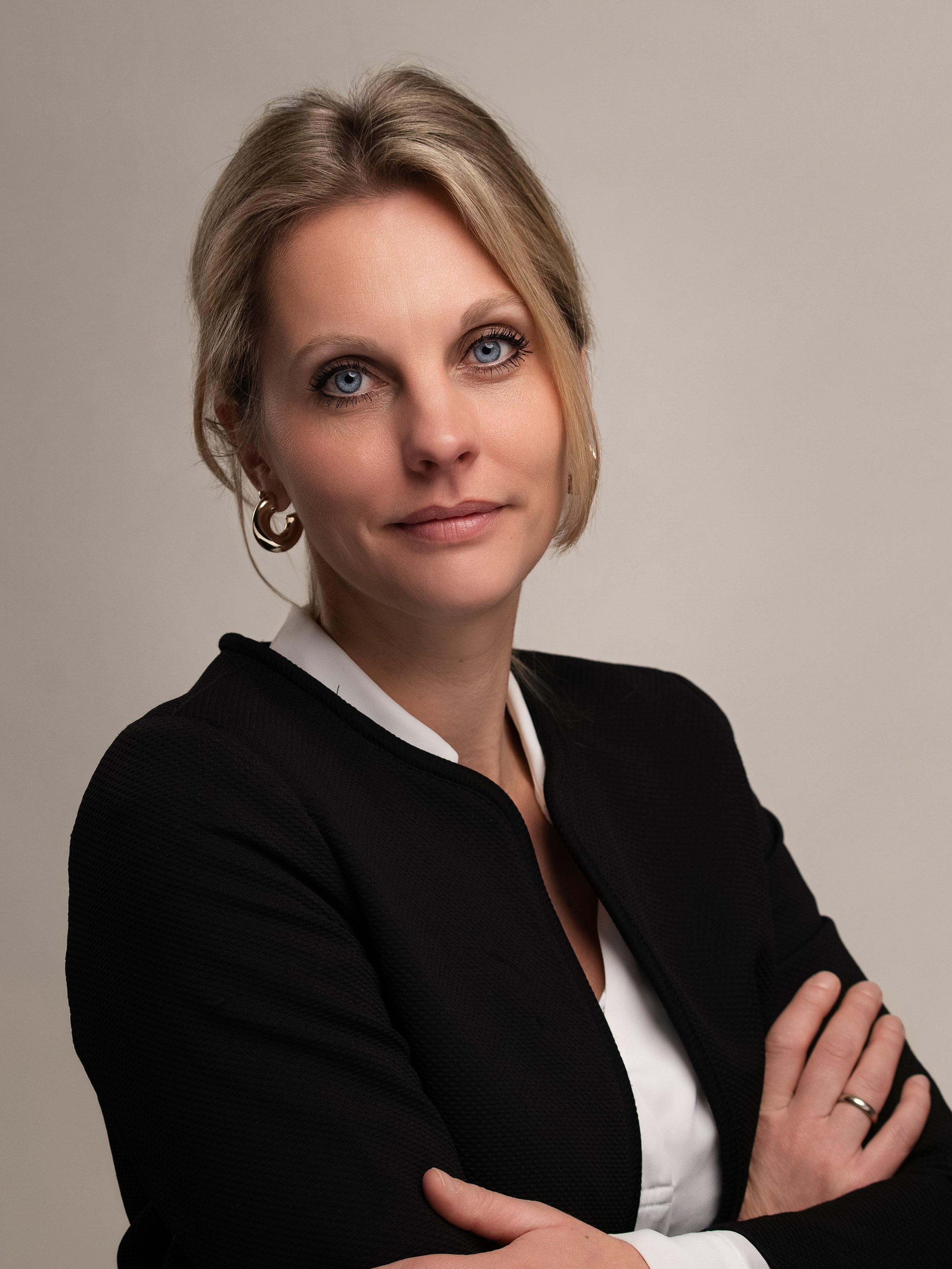 Portret van een vrouw met blonde haren, blauwe ogen en een lichte glimlach, gekleed in een zwarte jas en witte blouse, tegen een neutrale achtergrond.