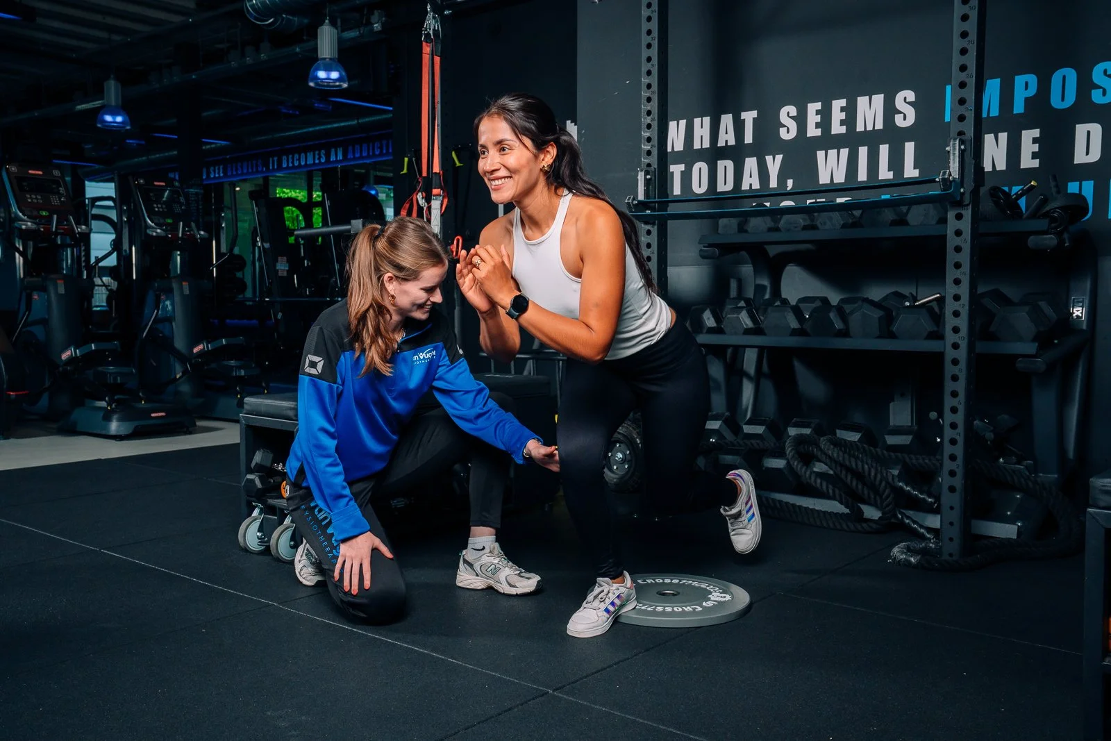 Vrouw die een oefening doet onder begeleiding van een personal trainer in een sportschool, terwijl ze op een plaatje staat met een opgeheven knie.