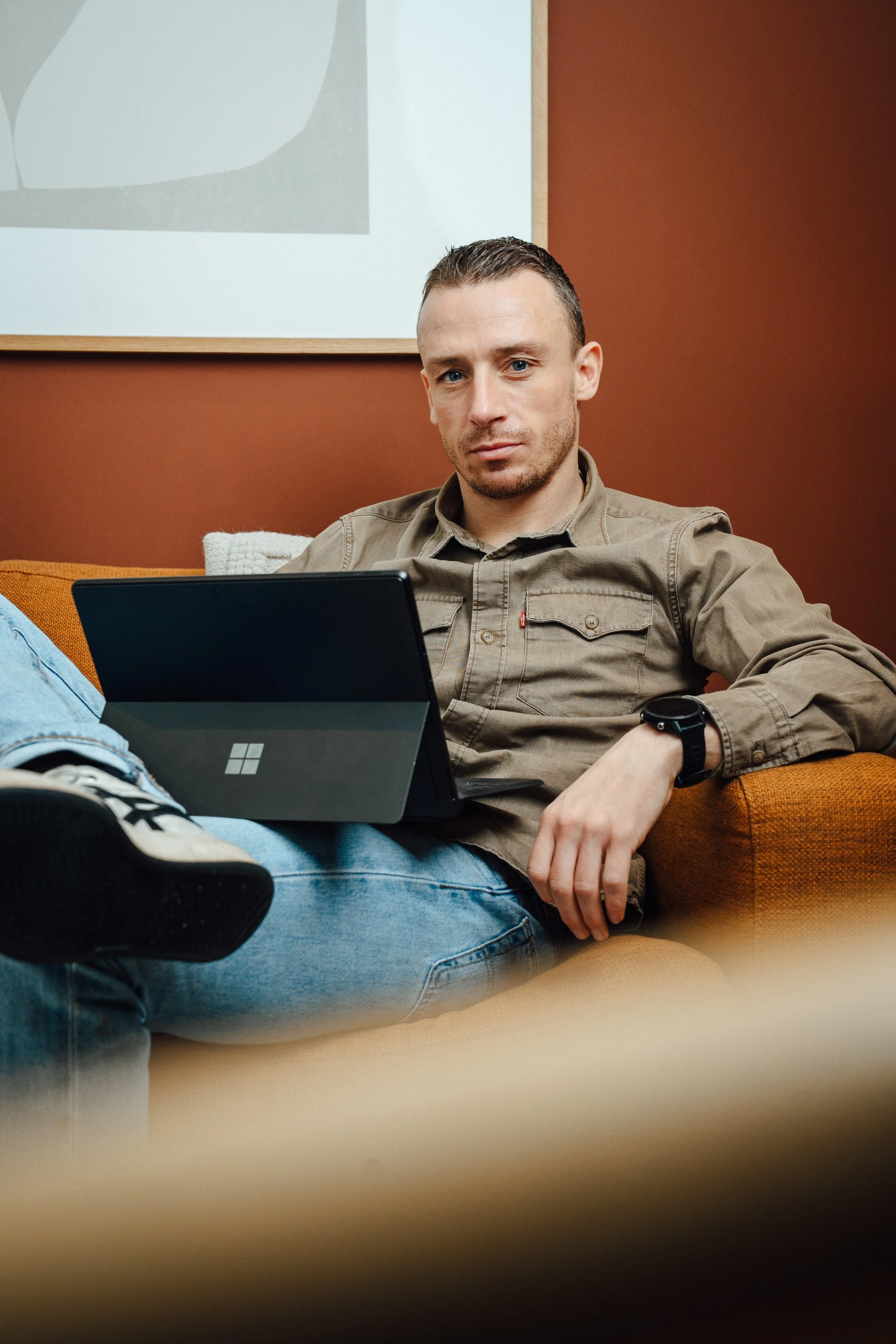 Een man zit op een bank met een laptop op zijn schoot, gekleed in een beige overhemd en blauwe jeans, in een interieur met bruin/oranje muren en een schilderij aan de muur.