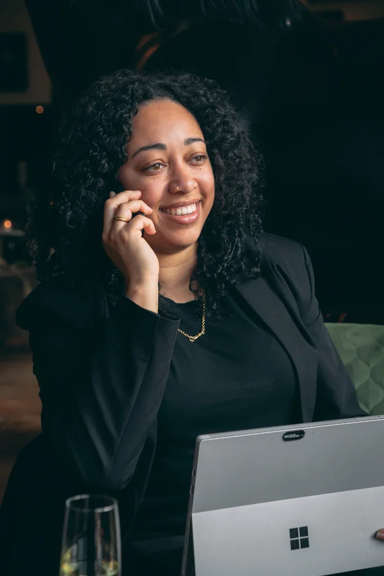 Vrouw met curly haar die aan de telefoon ligt, zittend achter een laptop met het logo van Microsoft, in een donkere omgeving.