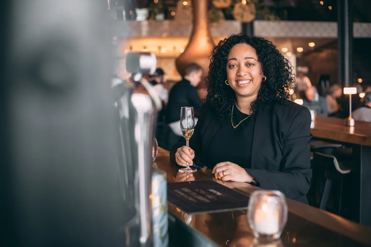 Een vrouw met krullend haar poseert met een glas champagne in een restaurant of bar, omringd door andere gasten en sfeervolle verlichting.