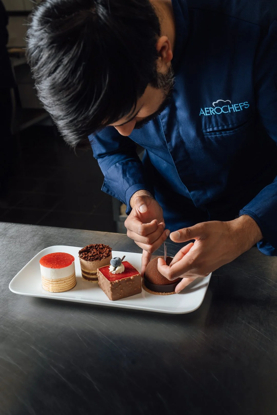 Een chef-kok die zorgvuldig een chocoladepraline voorbereidt, met vier verschillende verfijnde desserts op een wit dienblad voor zich.
