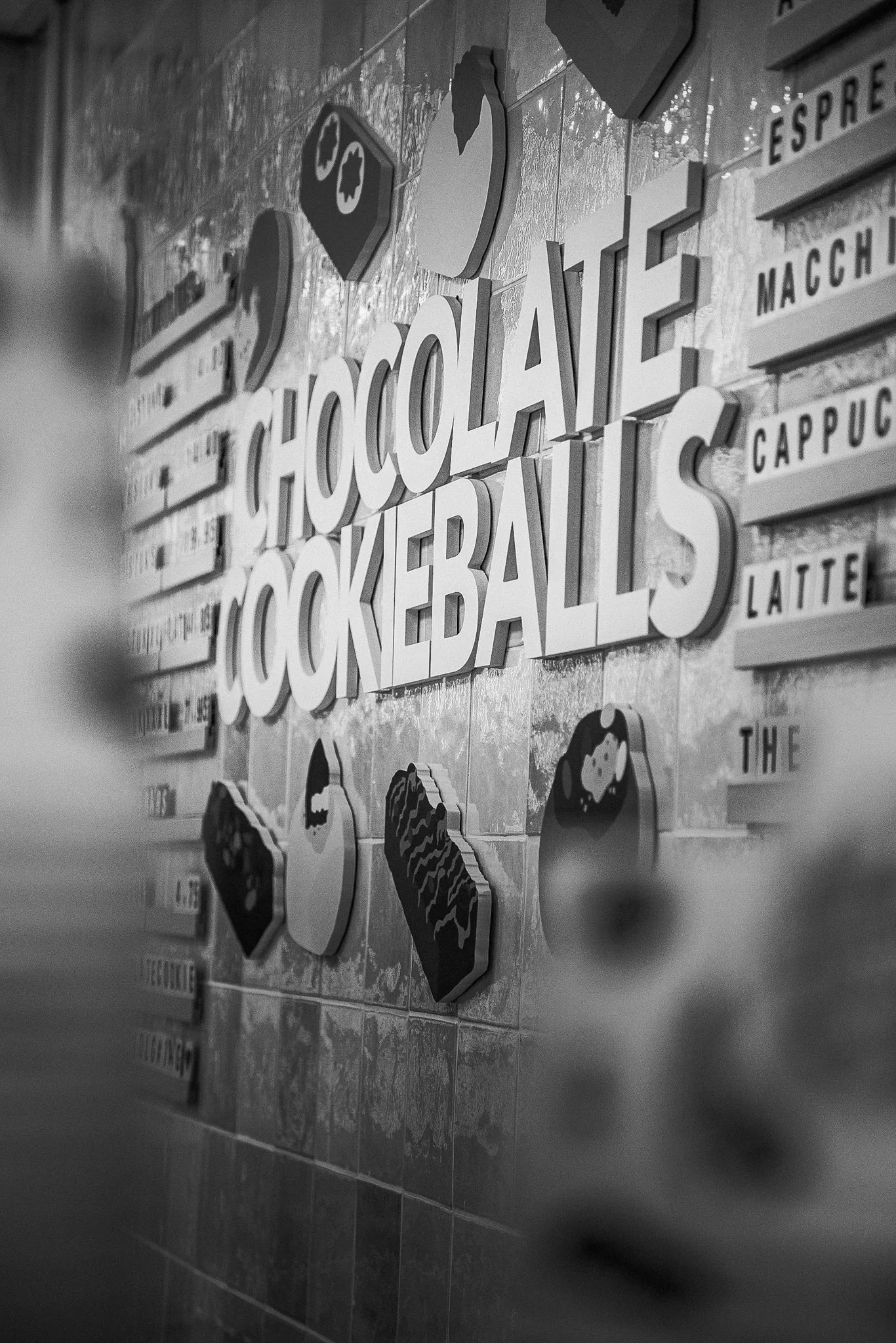 Zwart-wit foto van een muur met decoraties en tekst 'chocolate', 'cookie balls', en andere woorden, met illustraties van chocola, koekjes en latte op de muur.