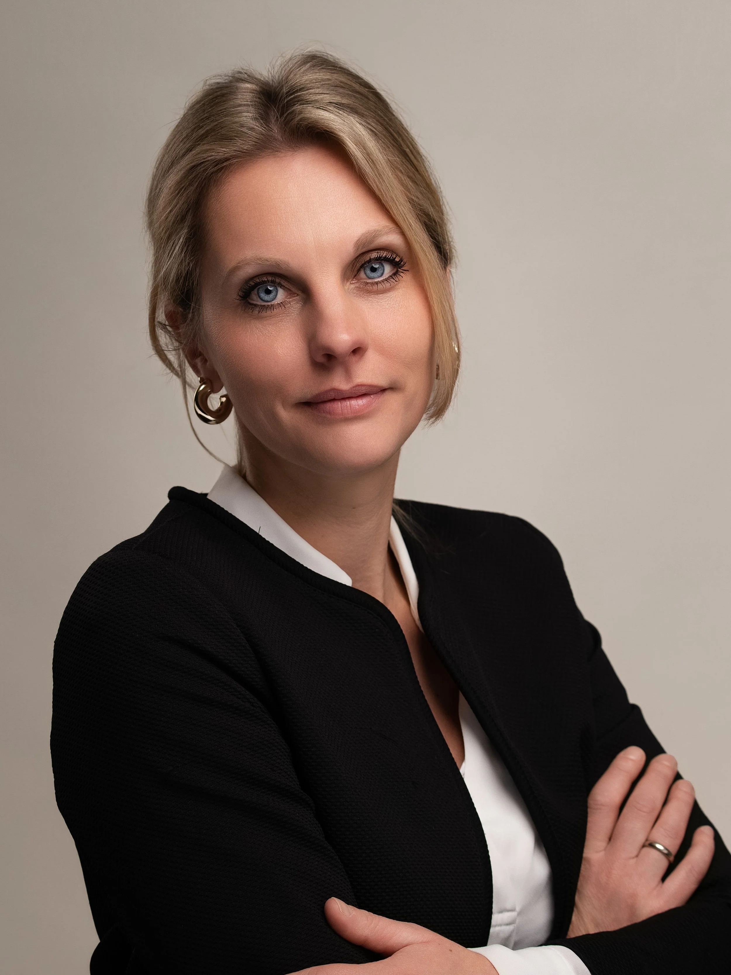 Portret van een vrouw met blond haar, blauwe ogen, draag een zwarte blazer en witte blouse, heeft haar handen gekruist, poseert voor een lichtgrijze achtergrond.