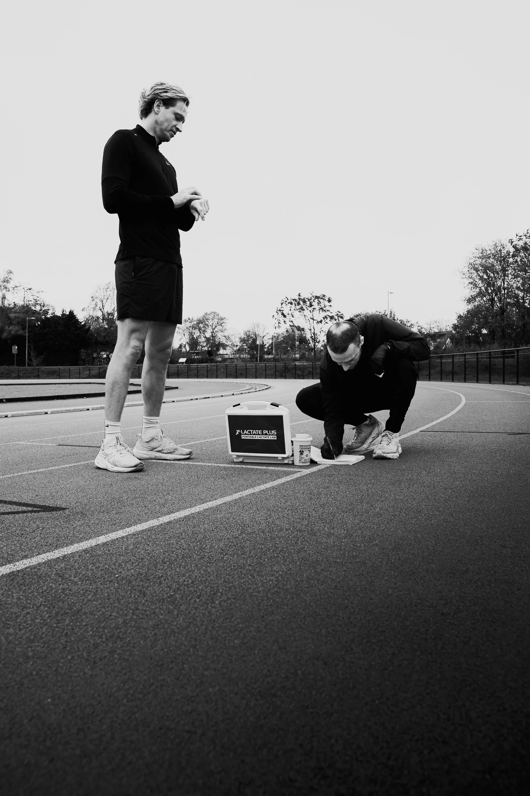 Twee mannen op een atletiekbaan, een van hen staat en kijkt naar de ander die op de grond zit en iets bekijkt, met een draagbare lactatentester en medicijnen in de buurt, in zwart-wit.